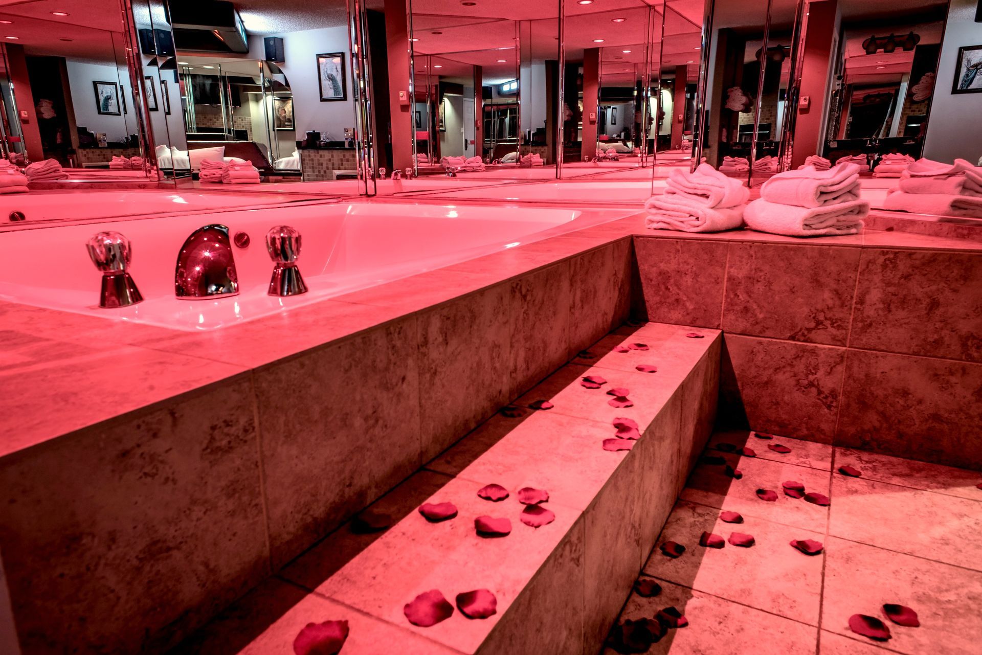 Romantic hotel suite with a red-lit jacuzzi, scattered rose petals, and multiple mirrors.