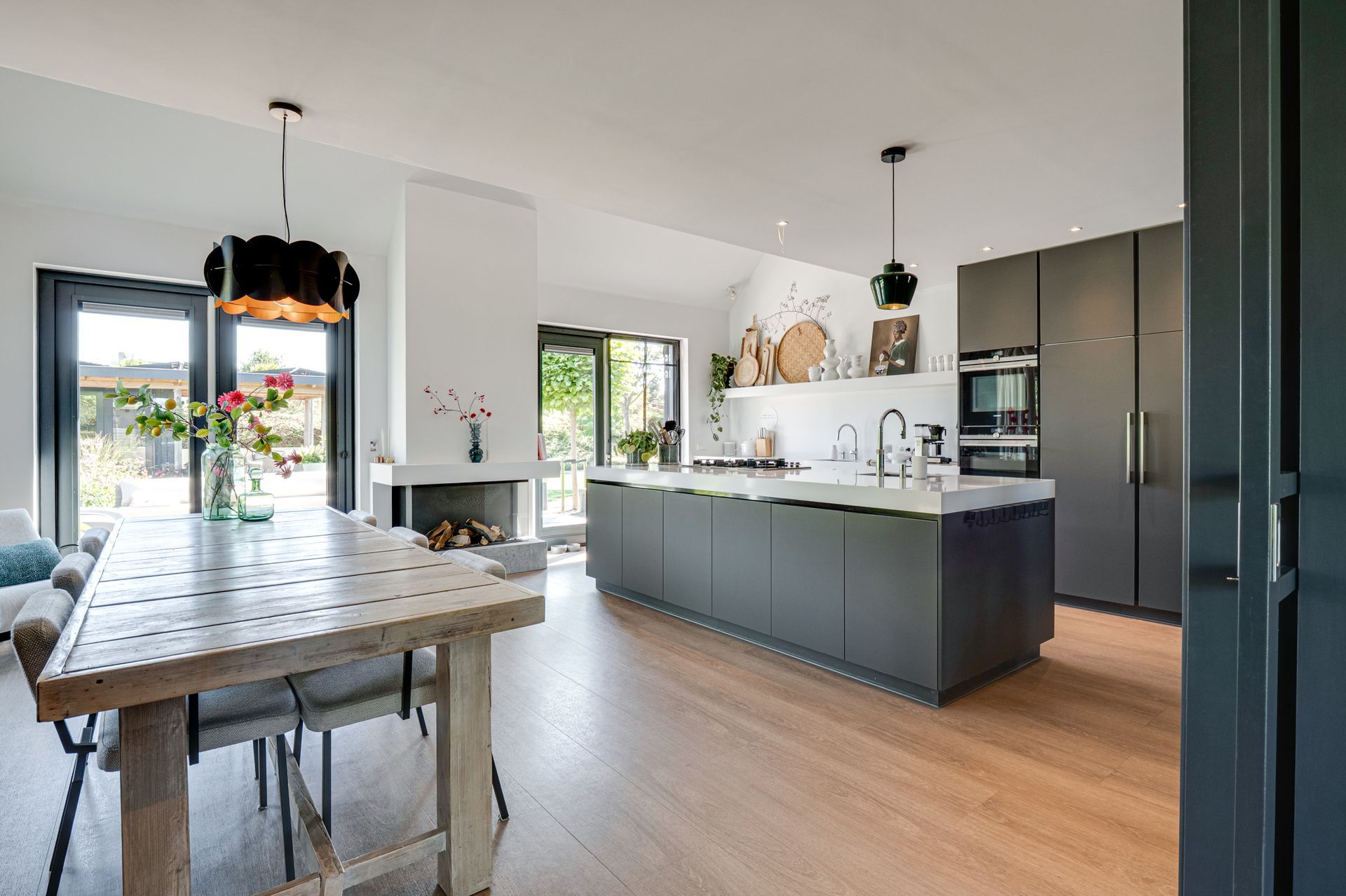 Moderne open keuken met grijs kookeiland, houten vloer en eettafel bij glazen schuifdeuren.