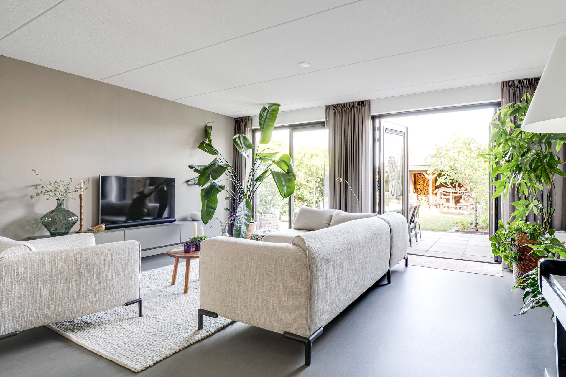 Lichte, moderne woonkamer met witte banken, tv, planten en schuifdeuren naar een terras.