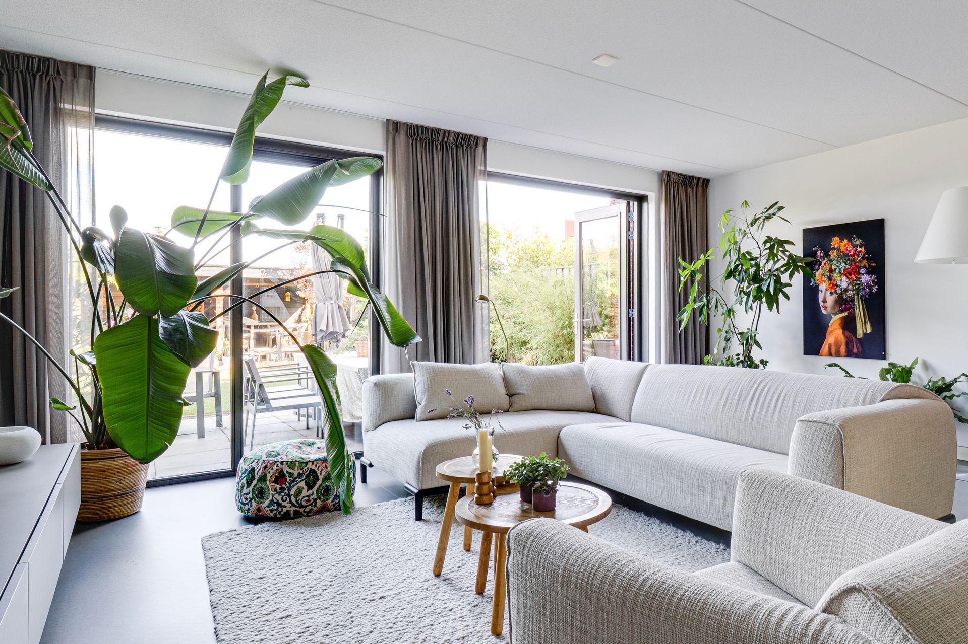 Lichte woonkamer met witte hoekbank, grote ramen, hoge planten en een kleurrijke muurschildering.