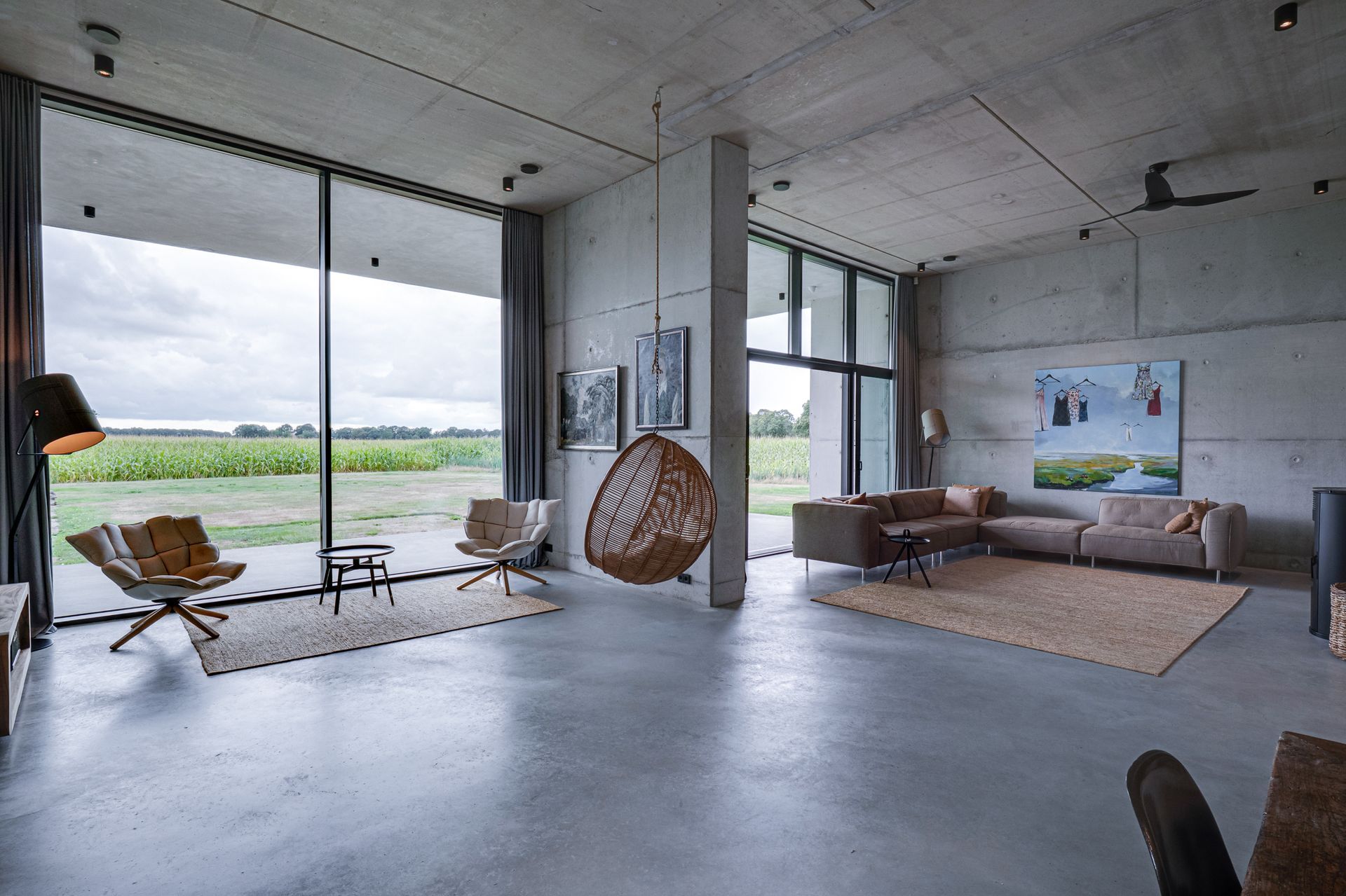 Moderne woonkamer met ramen van vloer tot plafond, grijze bank, rieten stoel en uitzicht op een open veld.