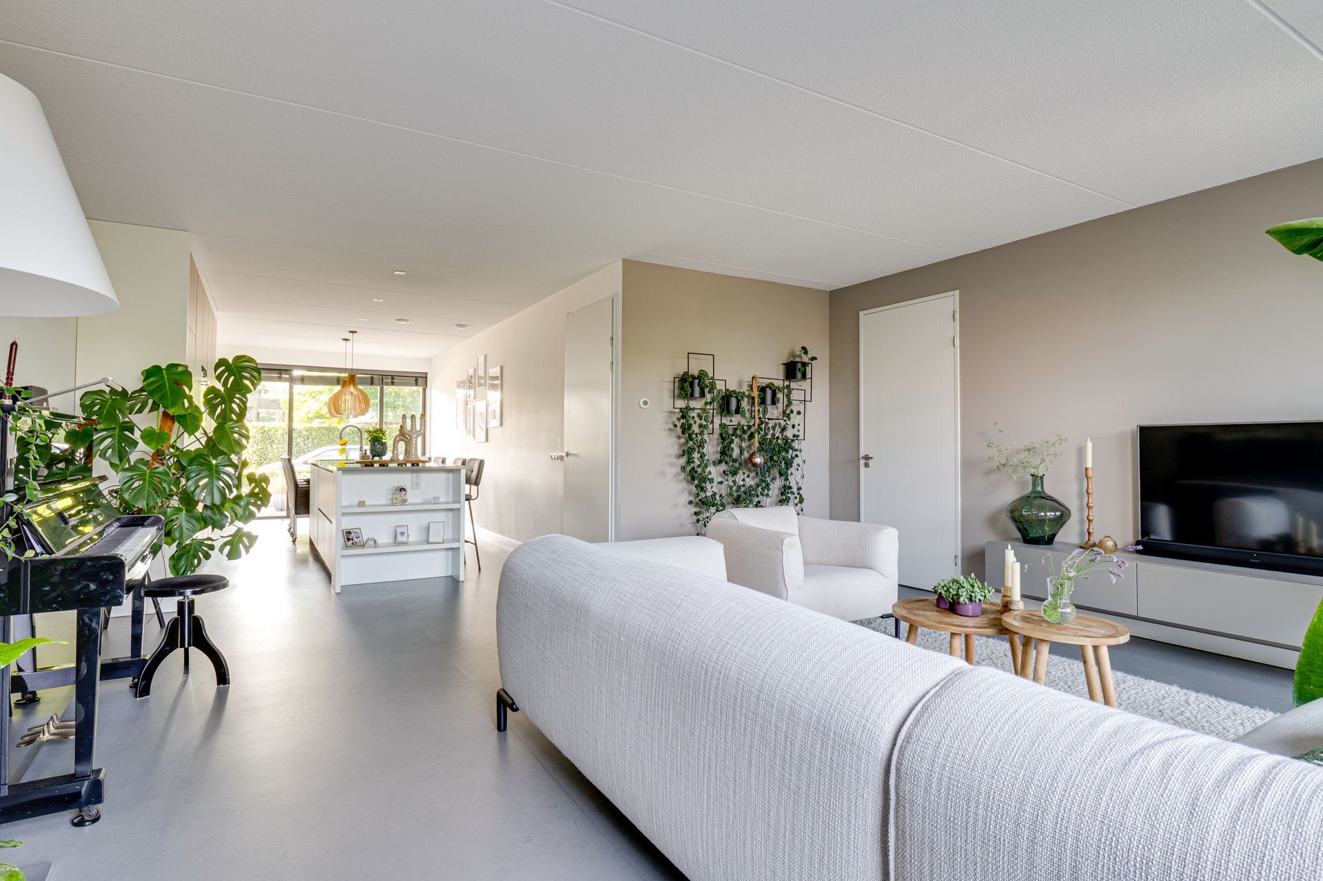 Lichte, moderne woonkamer met witte bank, tv, planten en een eethoek op de achtergrond.