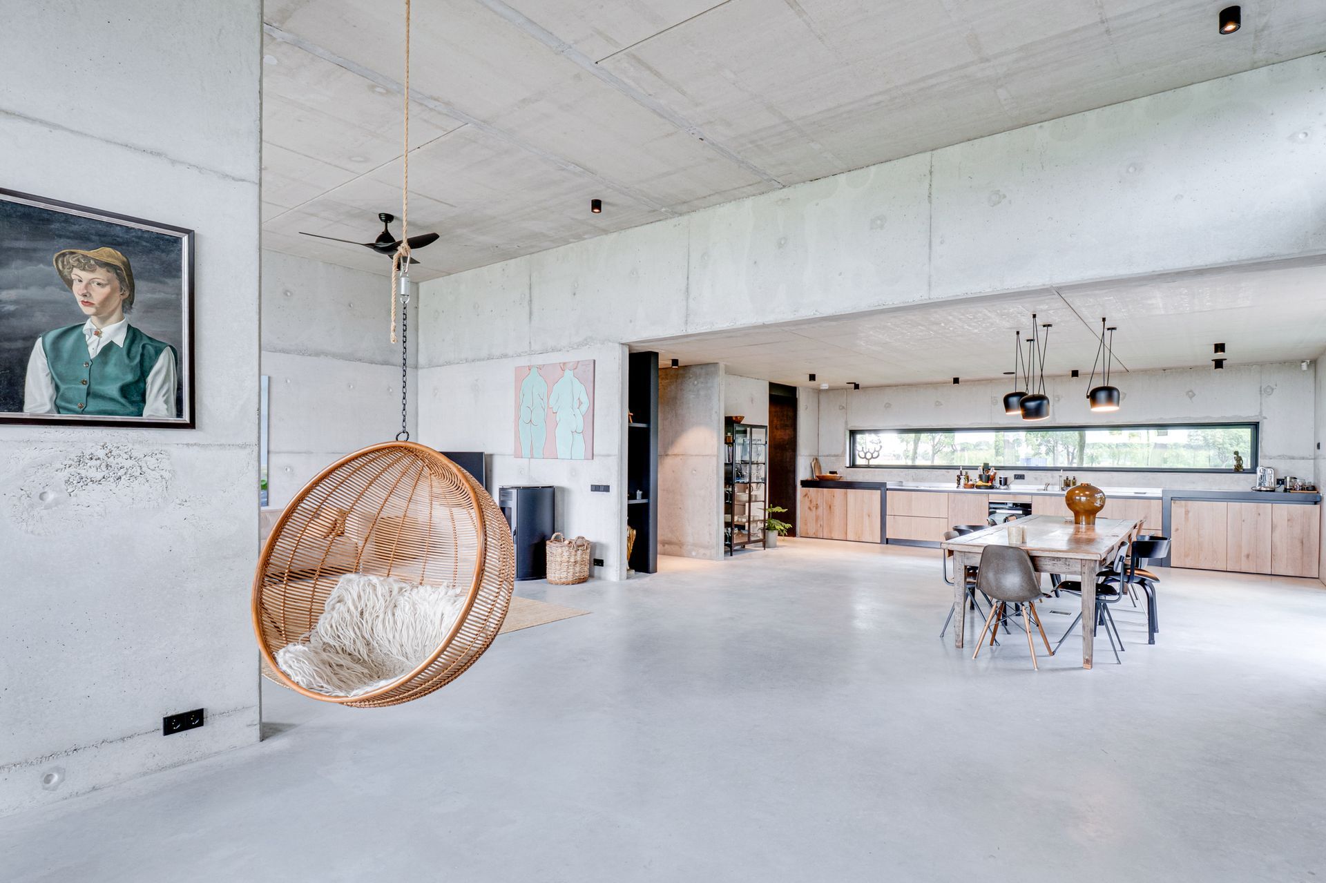 Ruime, onafgewerkte kamer met witte muren, rieten stoel, tafel en een groot raam.