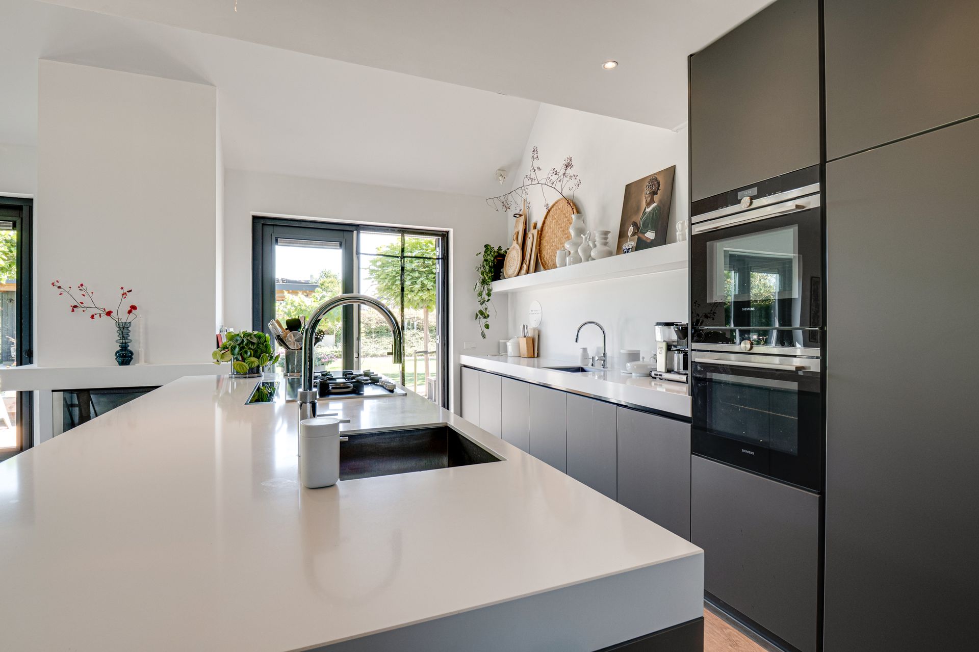 Moderne witte keuken met kookeiland, zwarte keukenkasten, ingebouwde oven en veel daglicht door grote ramen.