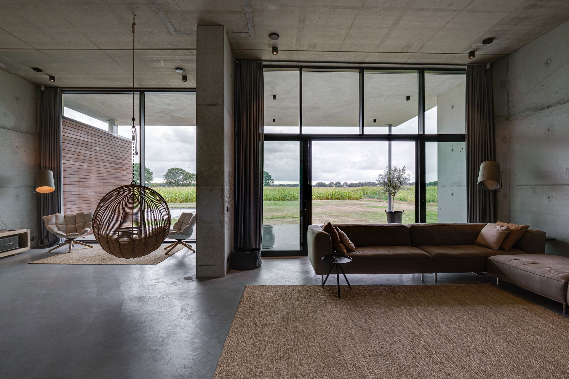Moderne betonnen woonkamer met bruine bank, hangstoel en grote glazen ramen met uitzicht op een groen veld.