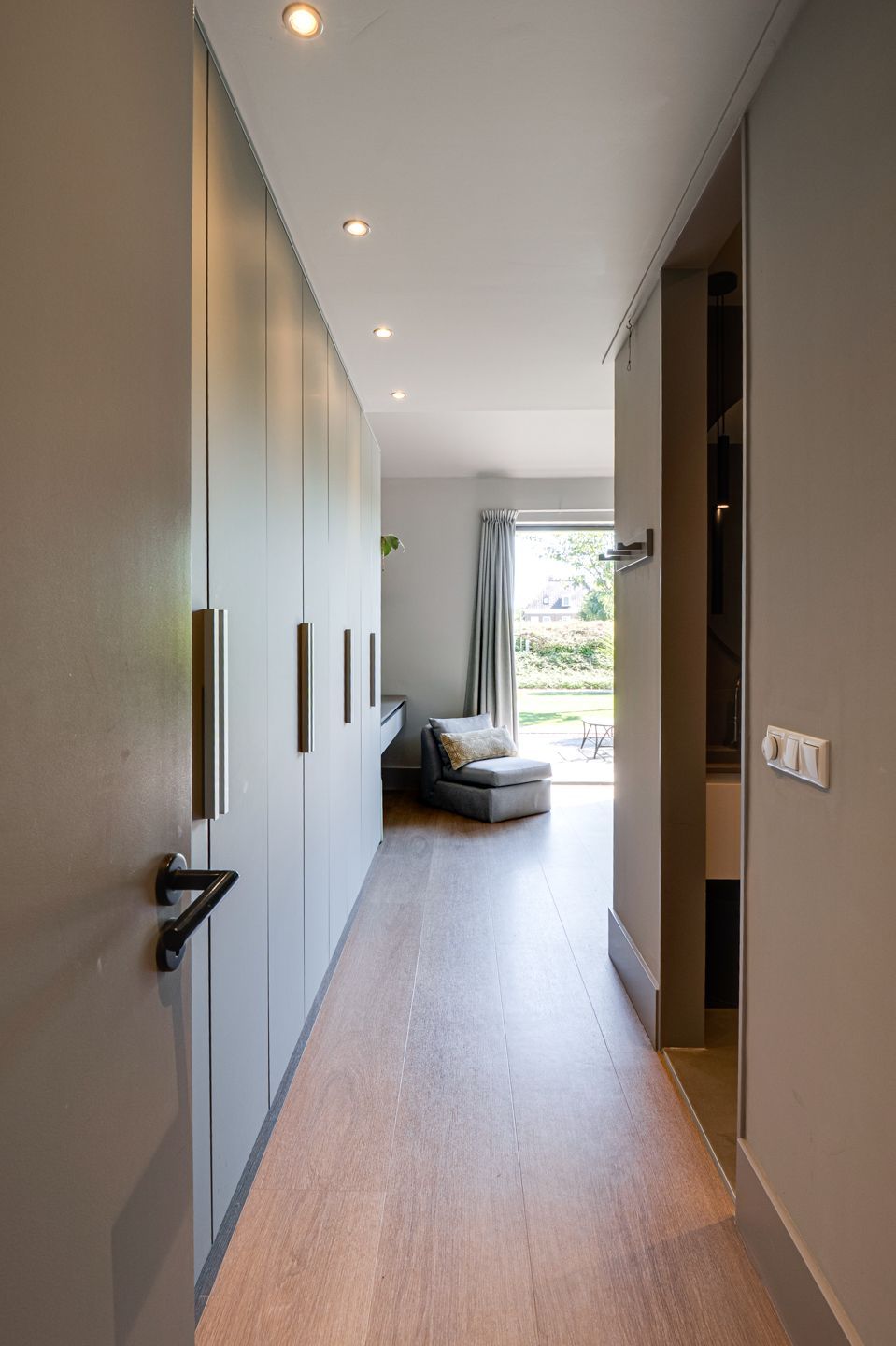 Smalle, moderne hal met houten vloer, inbouwspots, ingebouwde kasten en een stoel bij een licht raam.