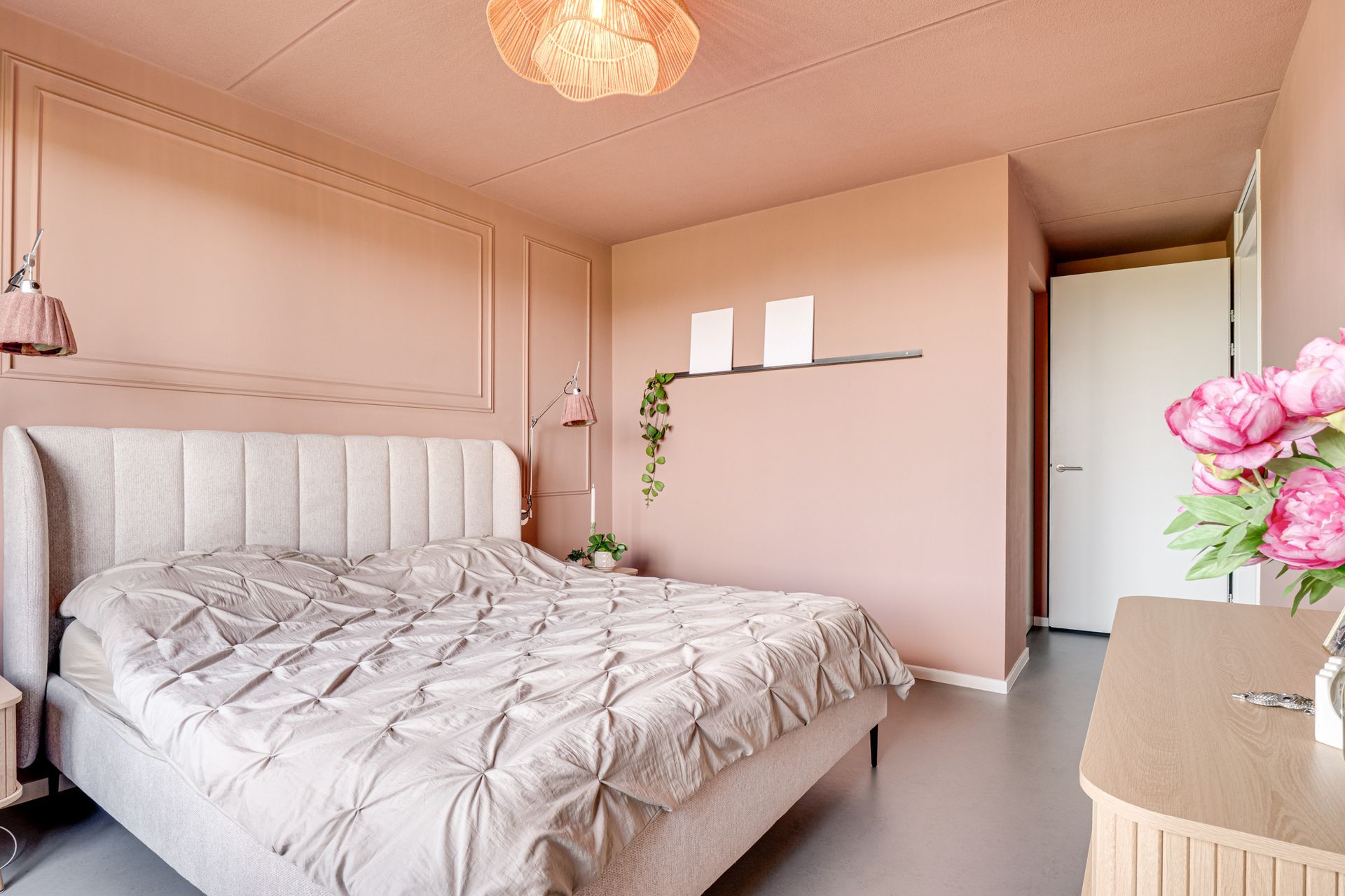 Roze slaapkamer met een wit bed, een gecapitonneerd hoofdeinde, een commode en bloemen bij een open deur.