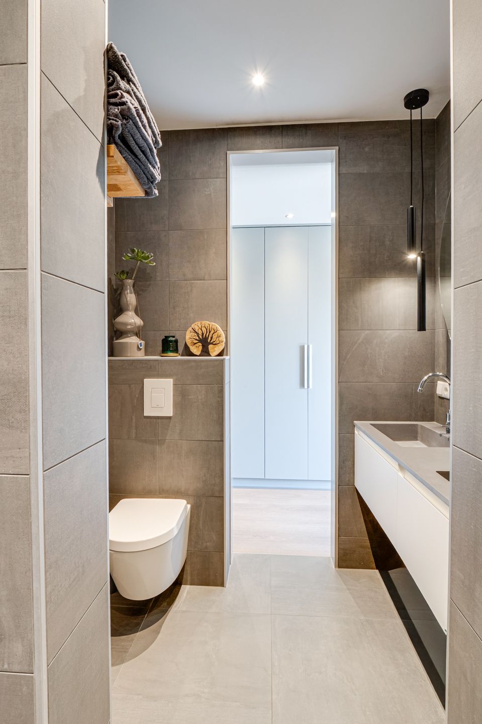 Moderne, smalle badkamer met grijze tegels, wandtoilet, badkuip en open doorgang naar een lichte hal.