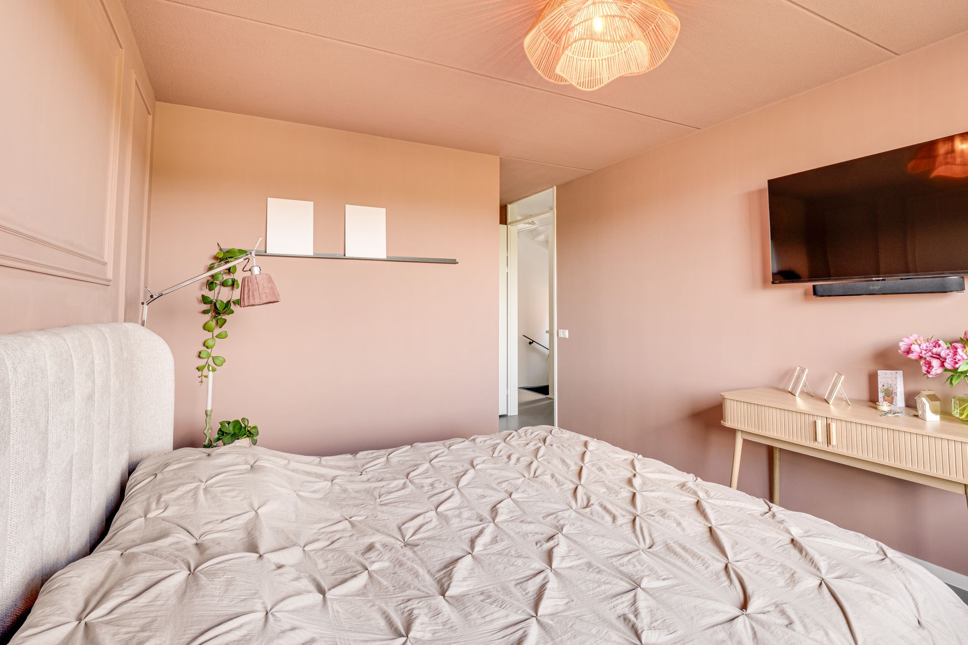 Gezellige slaapkamer met een wit bed, een aan de muur gemonteerde tv en een kaptafel in zachtroze tinten.