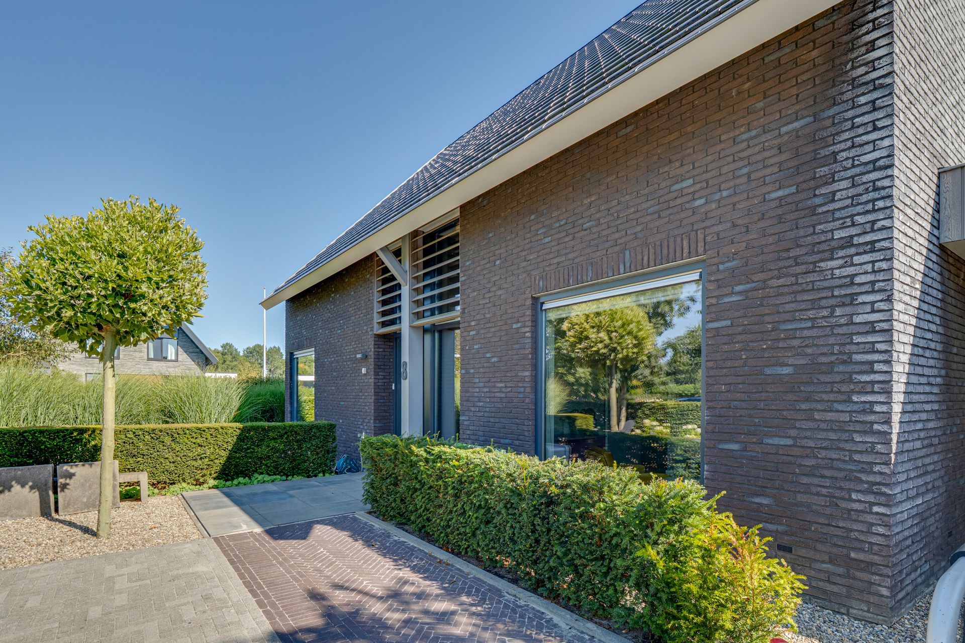 Moderne bakstenen woning met hagen, een kleine boom en een bestraat pad.
