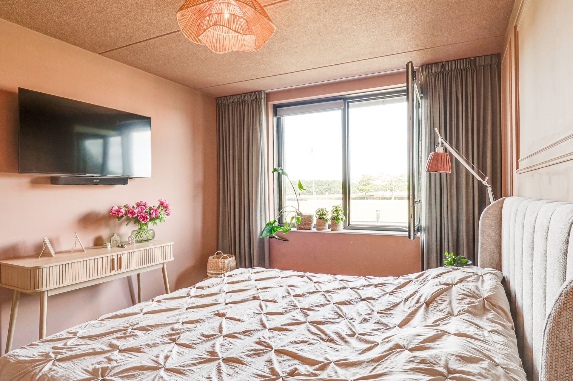 Lichte slaapkamer met een bed, aan de muur gemonteerde tv, raam en roze decoratie.