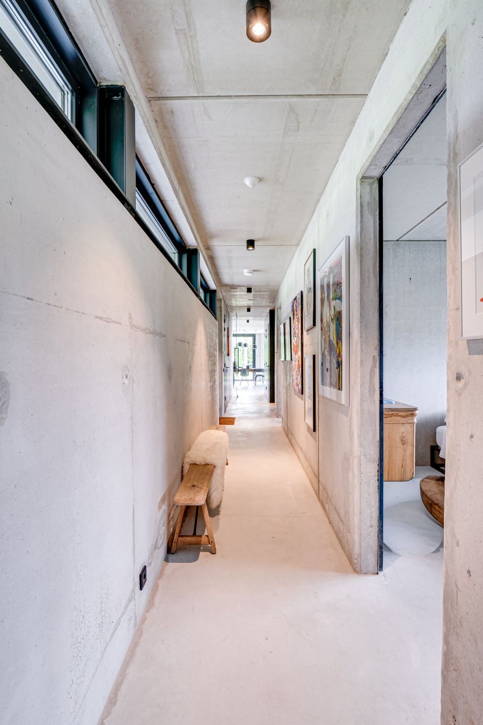 Lichte, smalle gang met witte muren, dakramen, ingelijste kunst en een klein houten bankje.