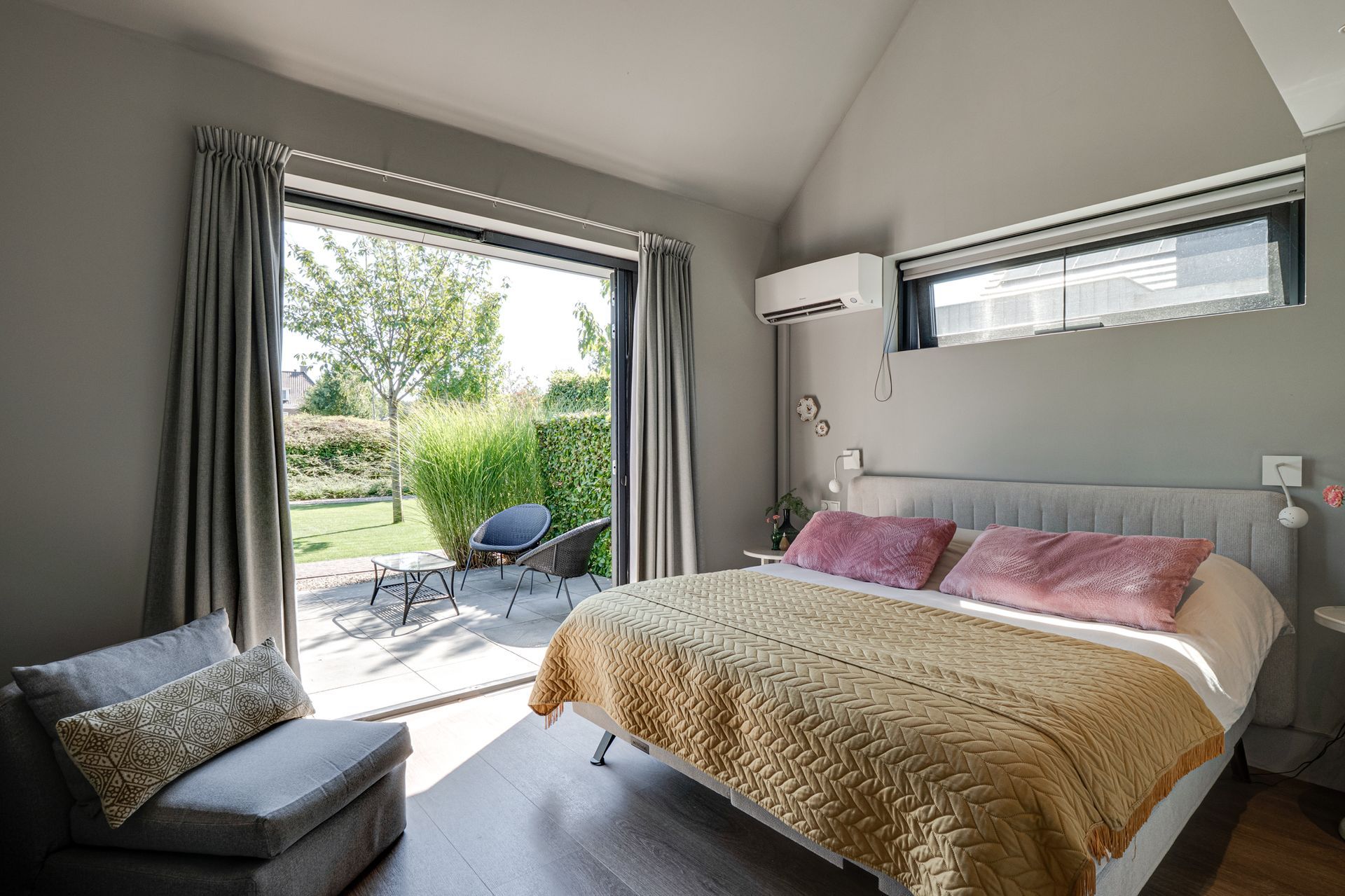 Lichte slaapkamer met openslaande terrasdeur, bed en grijze stoel met uitzicht op de tuin.