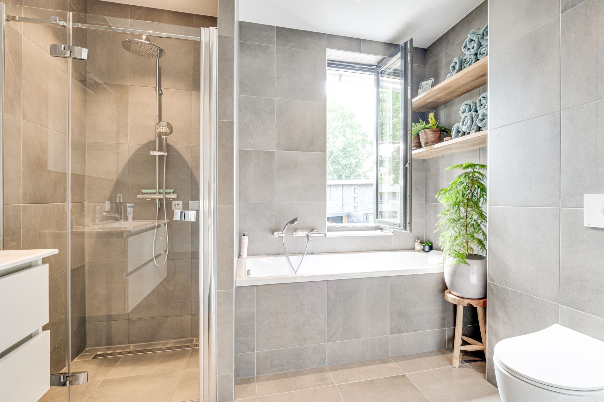 Moderne badkamer met glazen douche, bad bij het raam en planken met planten en toiletartikelen.