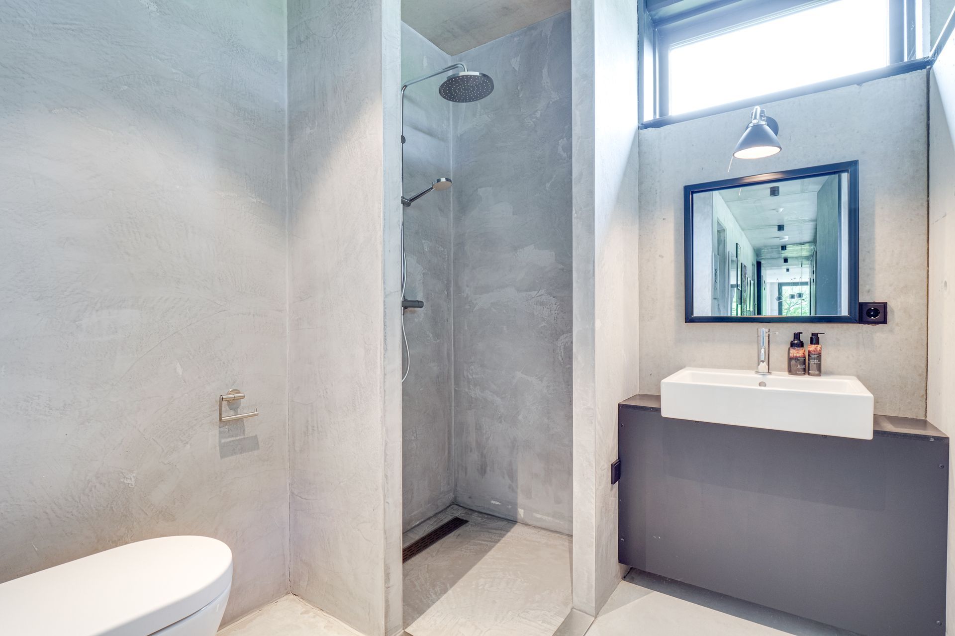 Moderne badkamer met glazen douchecabine, grijze wastafel, spiegel en groot raam dat veel licht binnenlaat.
