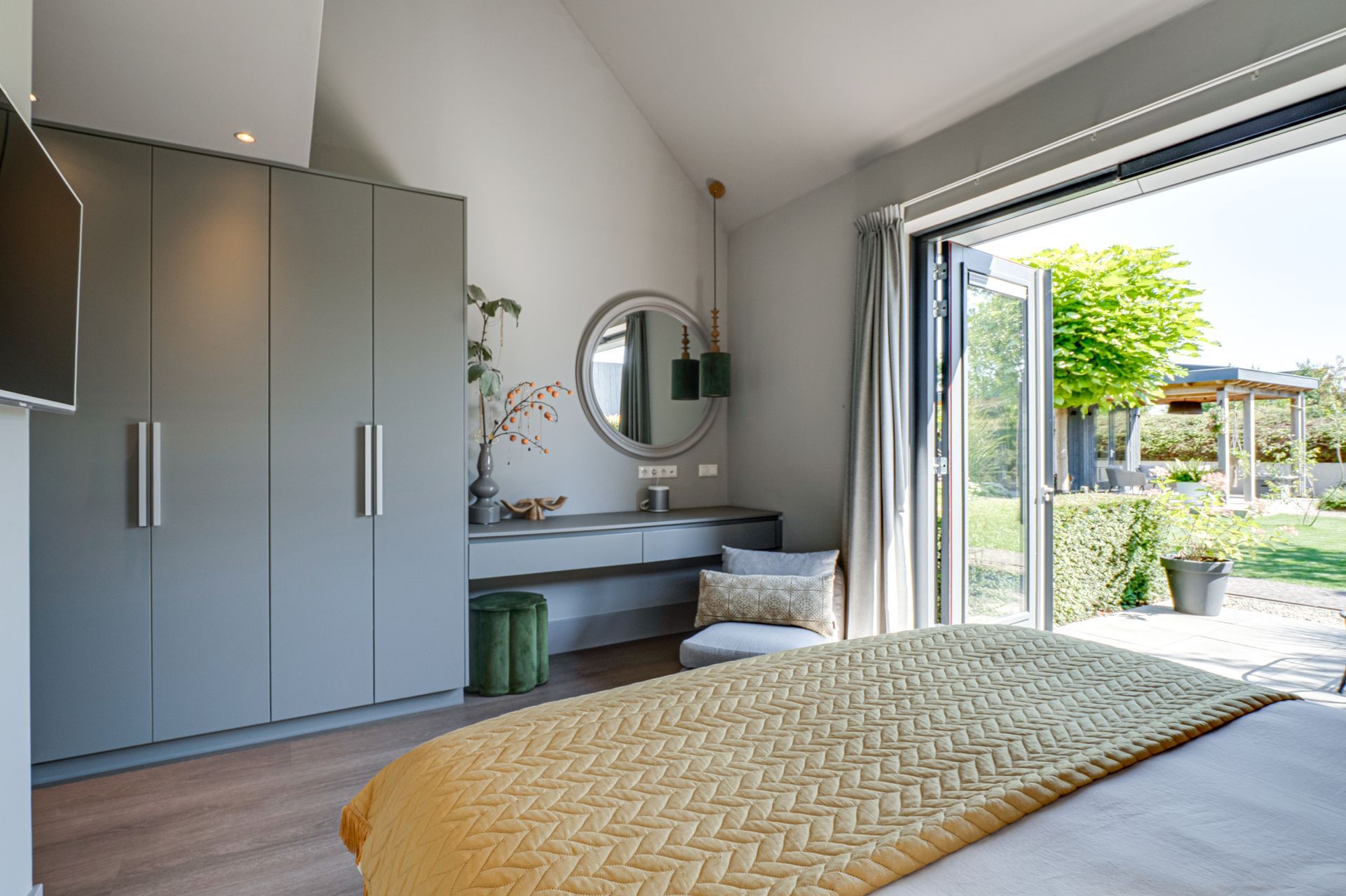 Moderne slaapkamer met grijze kledingkast, kaptafel, bed en openslaande glazen deuren naar een zonnig terras.