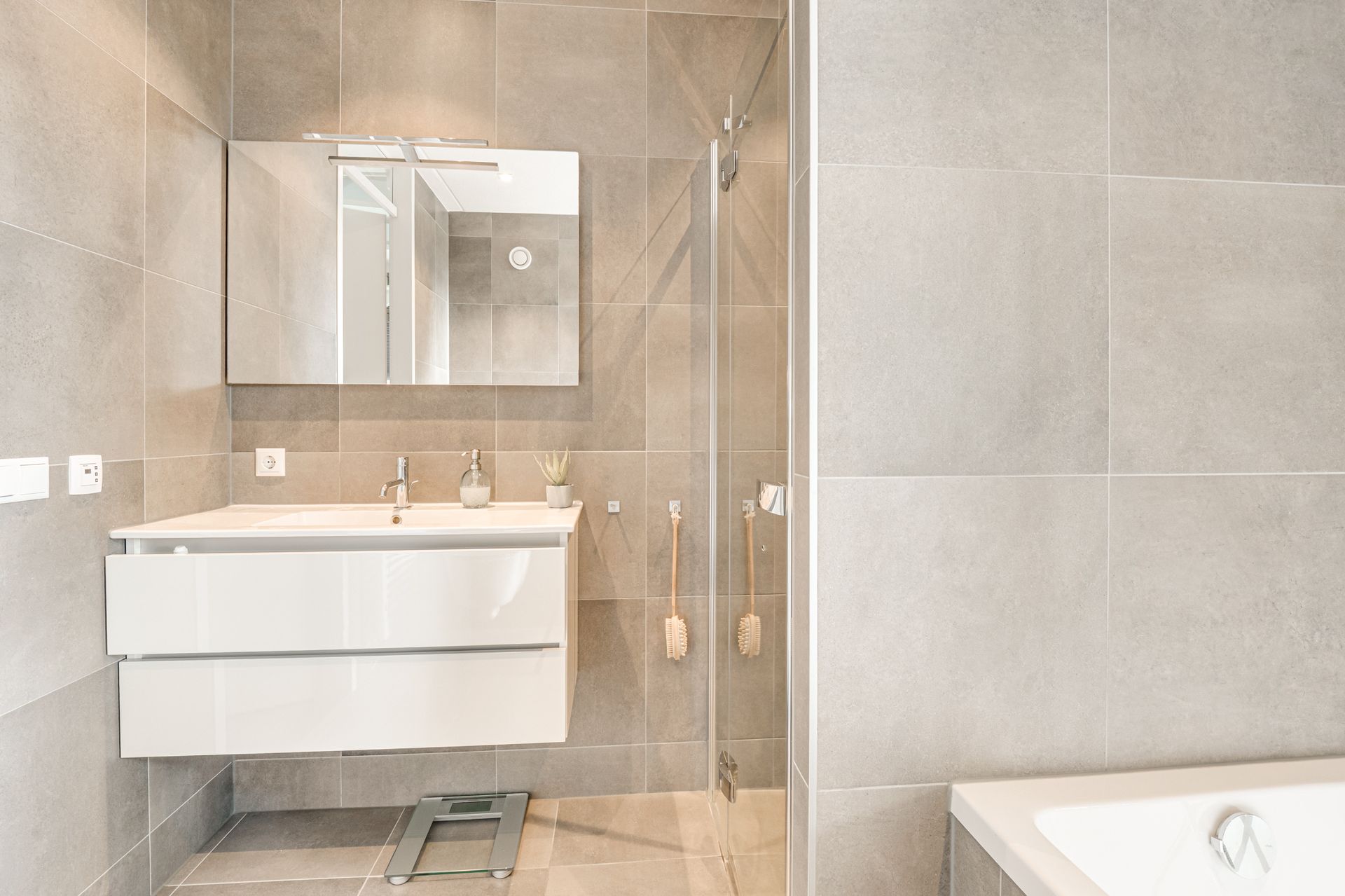 Moderne badkamer met wandgemonteerde wastafel, spiegel, glazen douchecabine en beige tegelwanden.