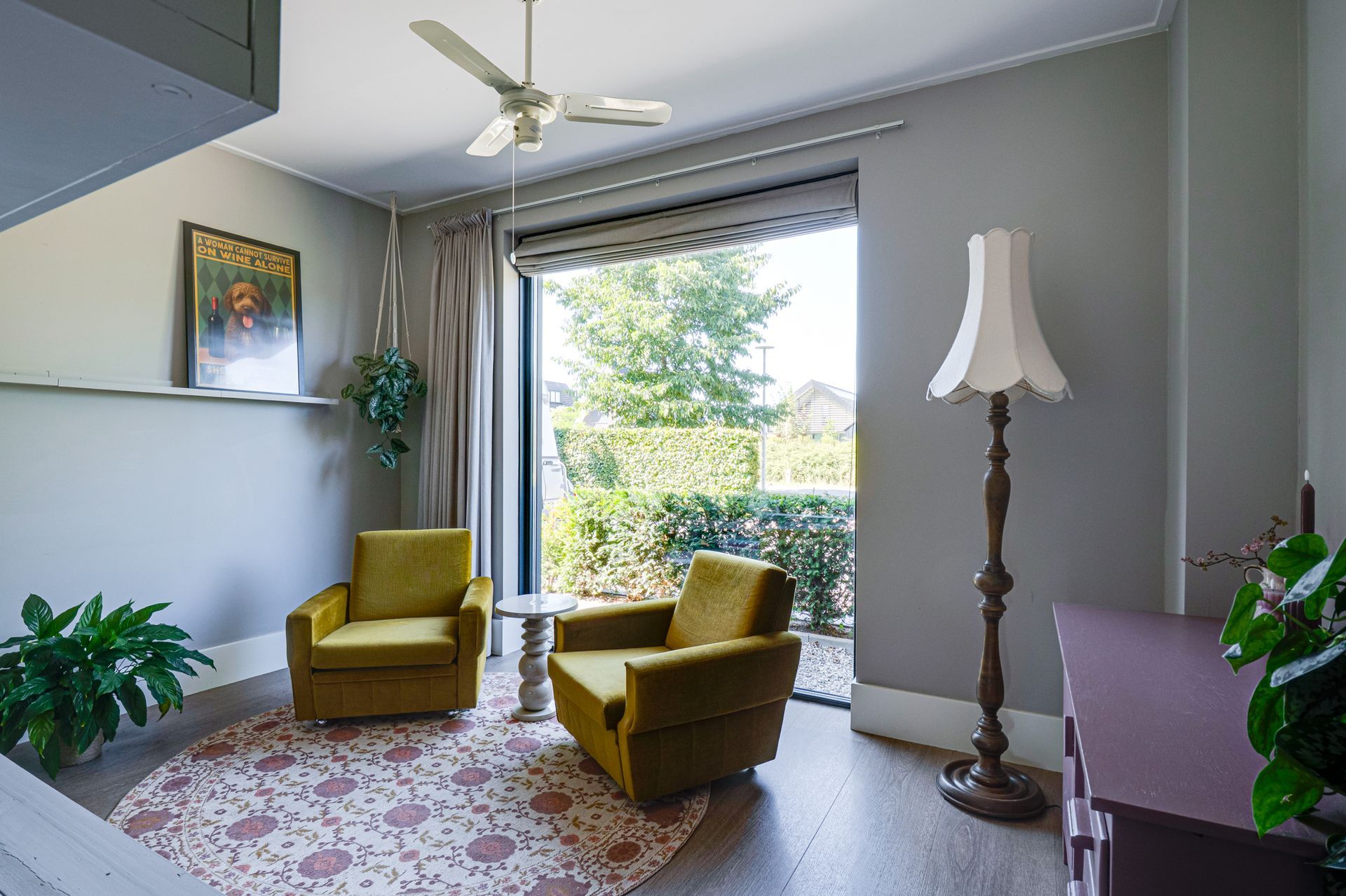 Lichte woonkamer met twee gele stoelen, groot raam, plafondventilator, staande lamp en een vloerkleed met patroon.