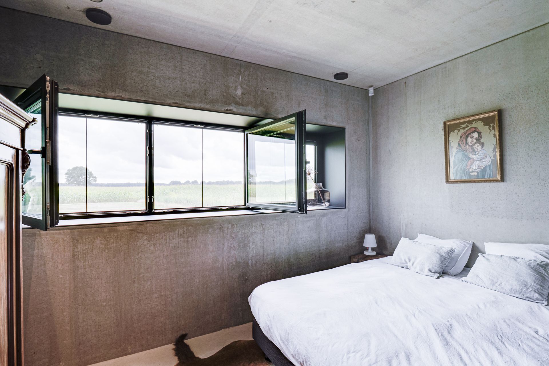 Lichte slaapkamer met grote ramen, een wit bed en grijze muren met uitzicht op een veld.