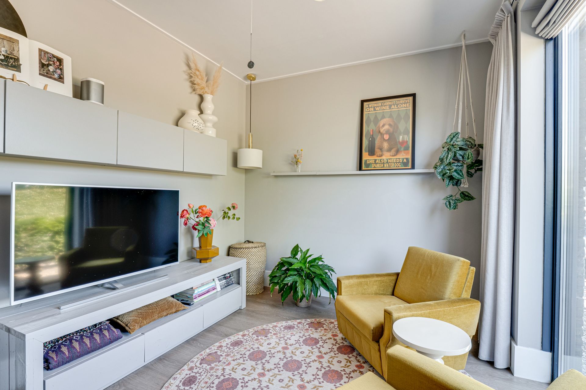 Moderne woonkamer met witte inbouwkasten, tv, mosterdkleurige fauteuil, potplanten en een groot raam met gordijnen.