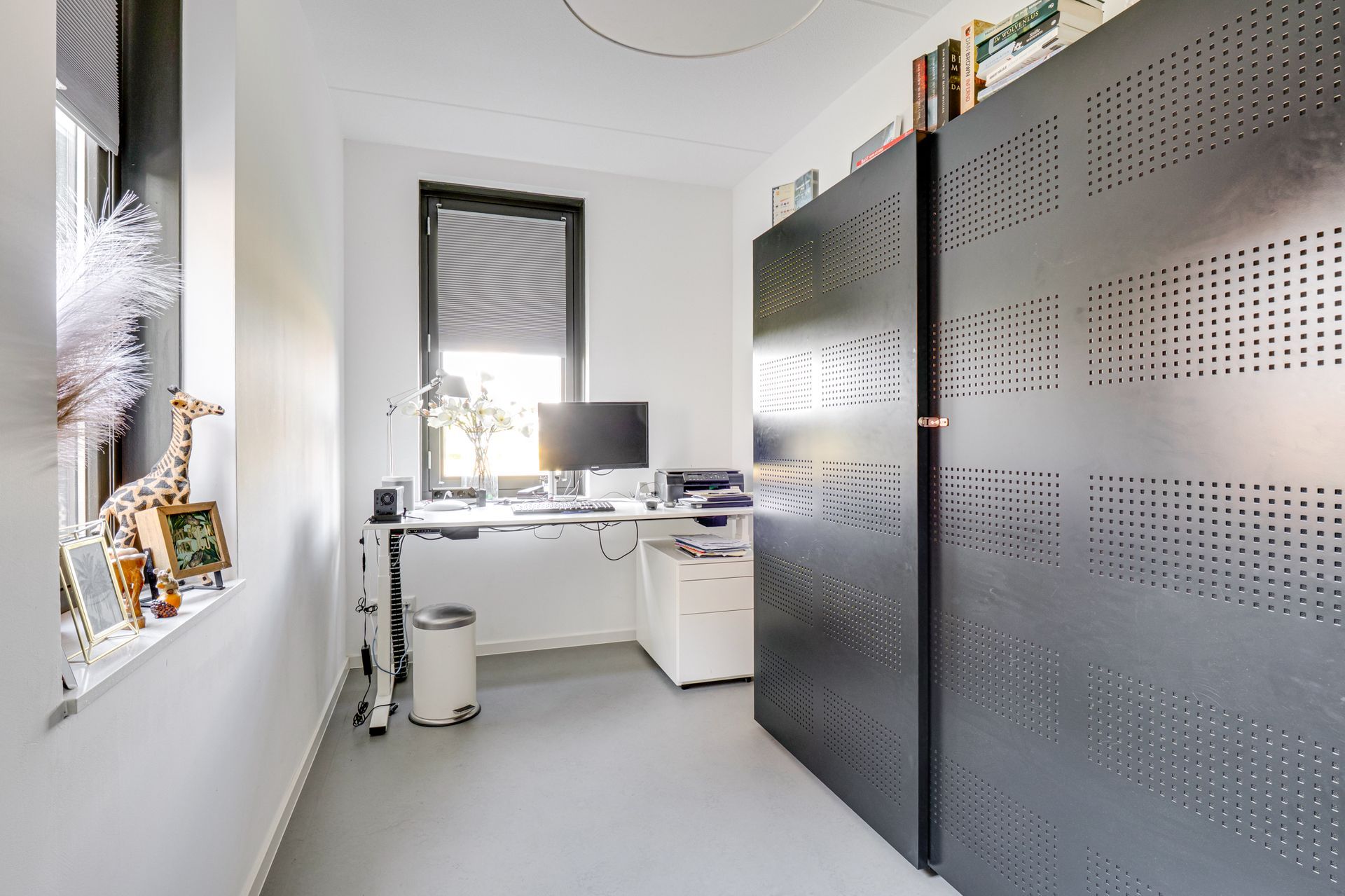 Een licht wit kantoor met een bureau, computer en donkere, geperforeerde opbergkasten langs de muur.