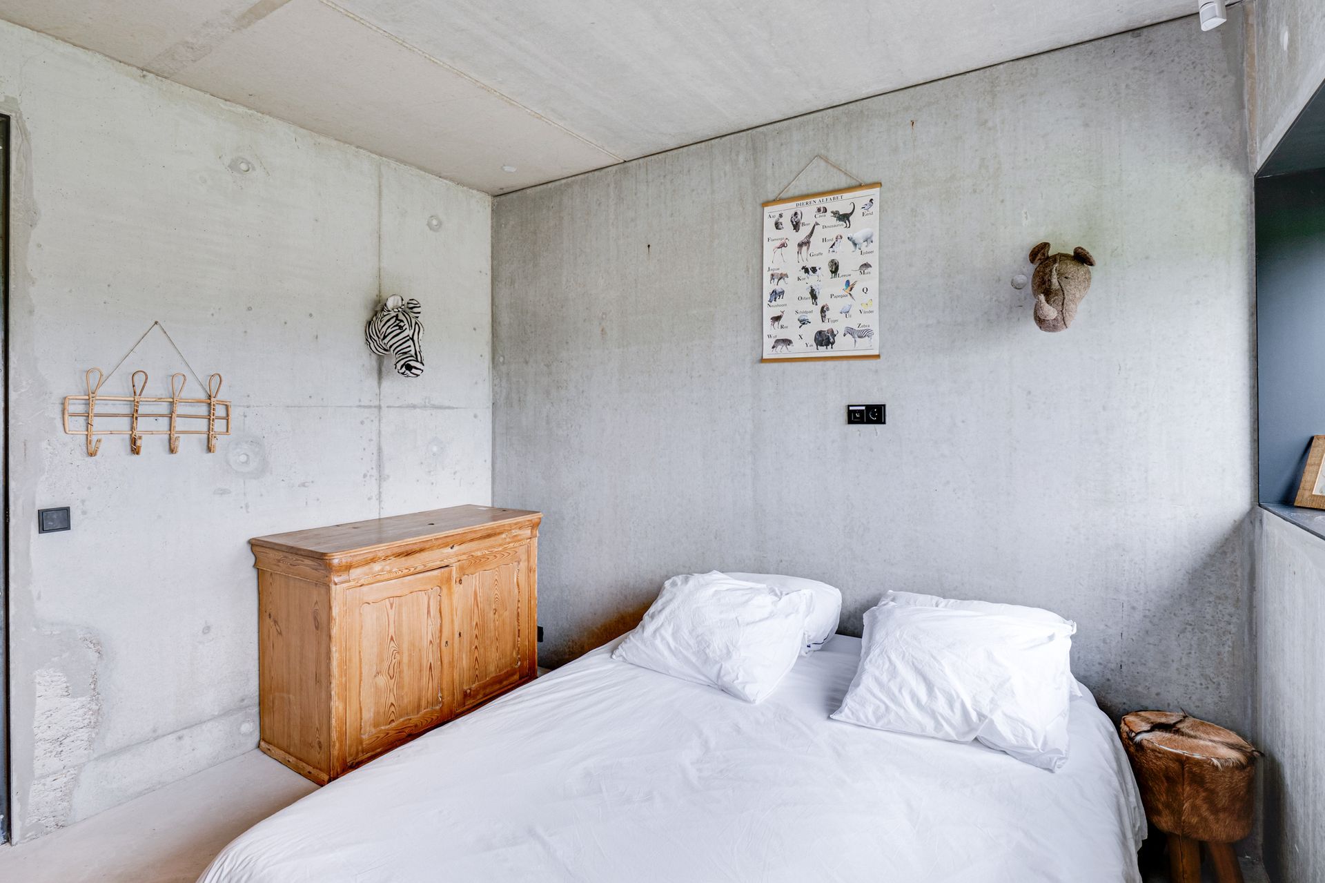 Kleine witte slaapkamer met een bed, houten commode, wandhaken en ingelijste decoratie.