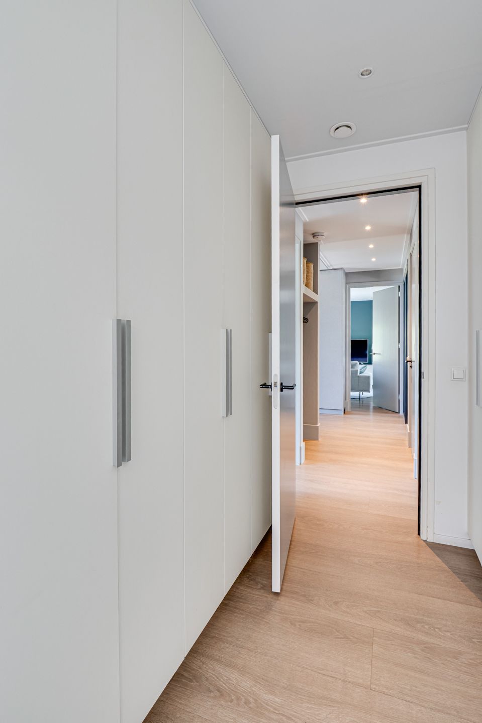 Lichte hal met witte inbouwkasten, open deuropeningen en een lichte houten vloer die toegang geeft tot een kamer.