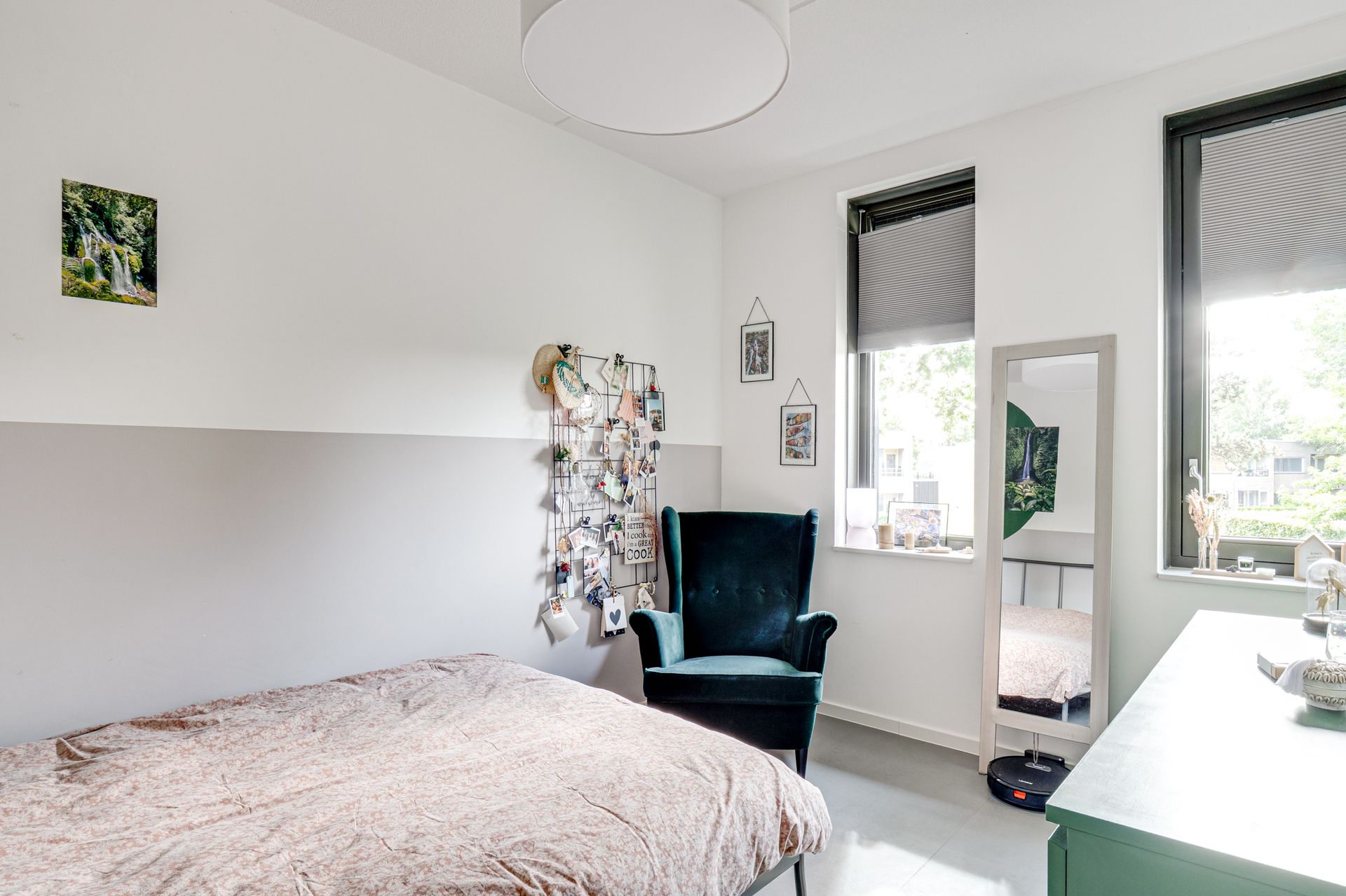 Lichte slaapkamer met een bed, zwarte stoel, witte muren, twee ramen en een staande spiegel.