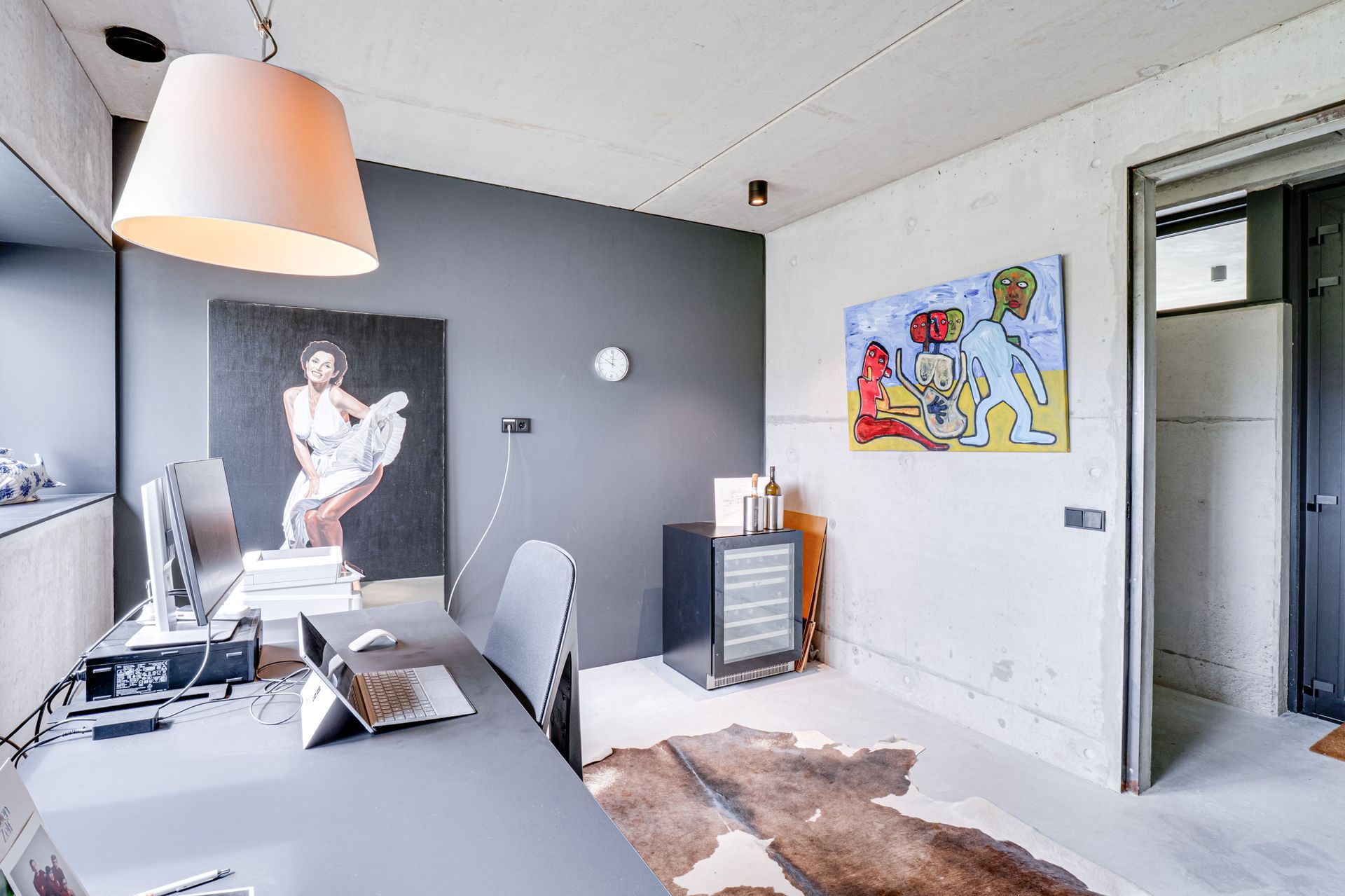 Lichte thuiswerkplek met wit bureau, grijze muurdecoratie, kleurrijk schilderij en open deur naar de gang.
