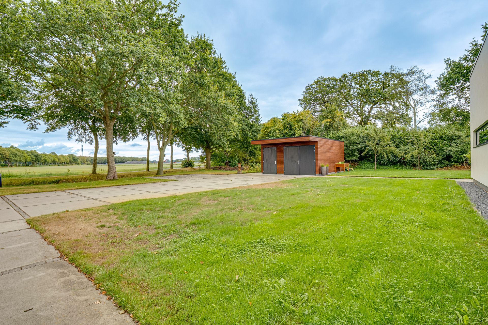 Een grasveld met een oprit, een klein schuurtje en bomen onder een heldere hemel.