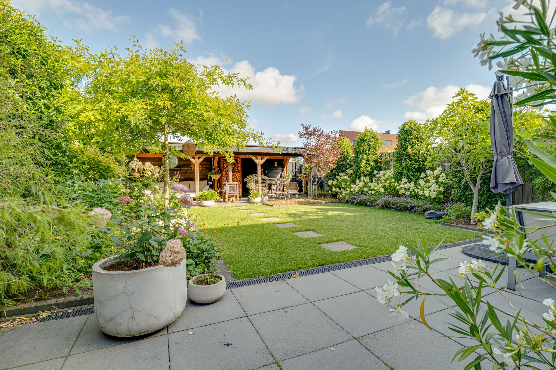 Zonnige achtertuin met terras, weelderig gazon en een houten pergola onder een blauwe hemel.