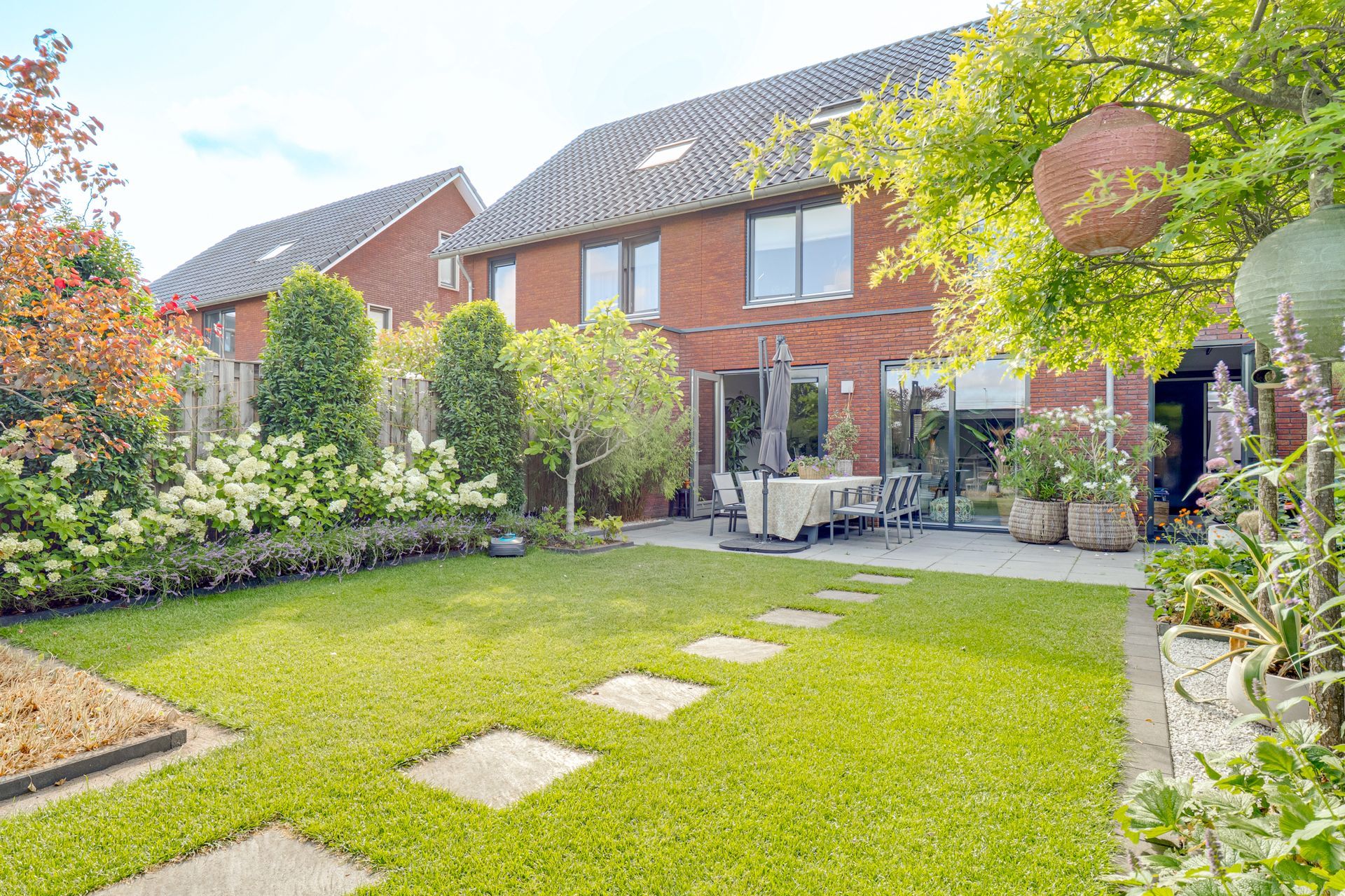 Achtertuin met groen gazon, stapstenen, terrasmeubilair en bakstenen rijtjeshuizen op de achtergrond.