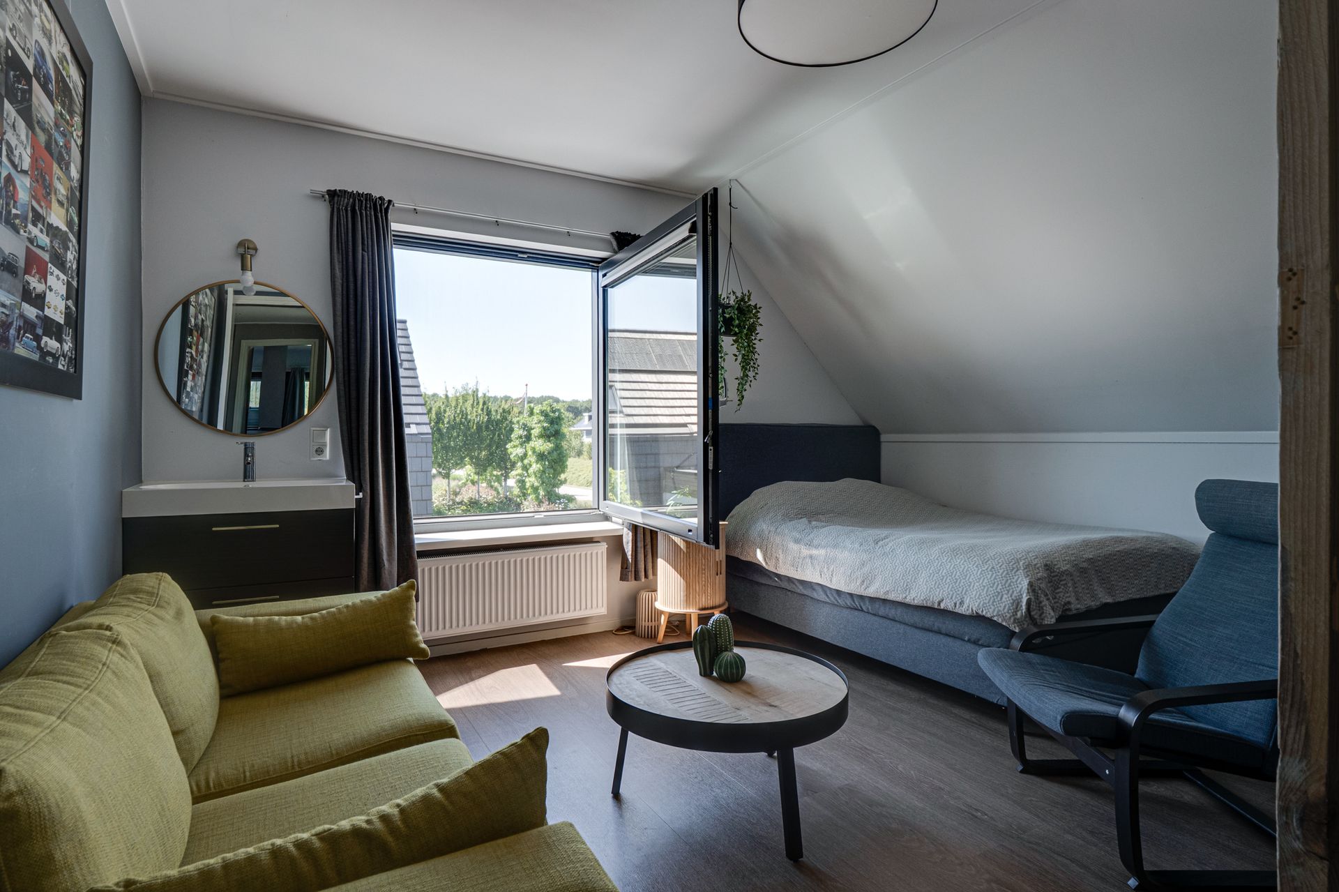 Lichte slaapkamer met een bed, groene bank, ronde tafel en een open raam met gordijnen.