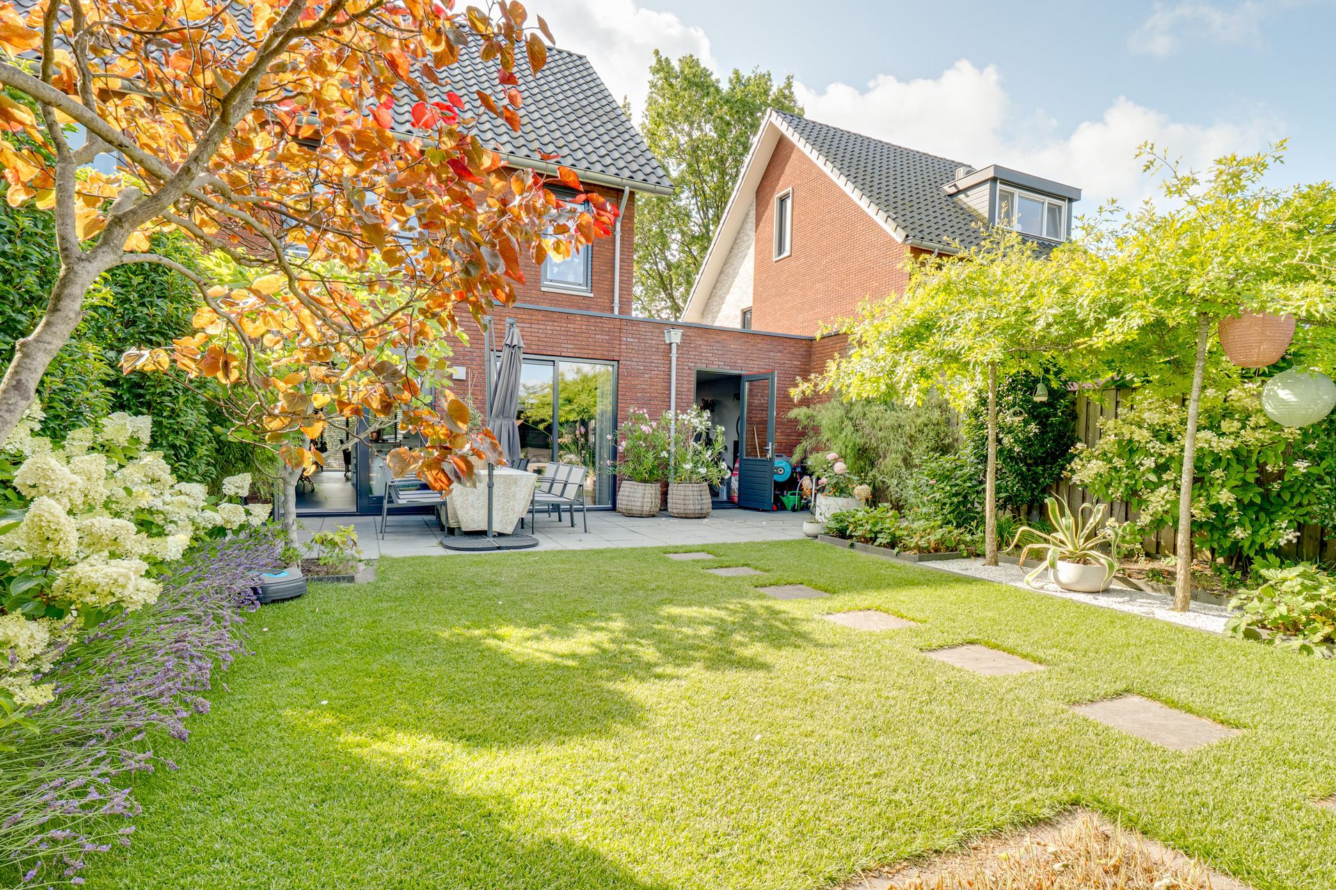 Zonnige achtertuin met groen gazon, bakstenen huisjes, bomen en terrasstoelen.