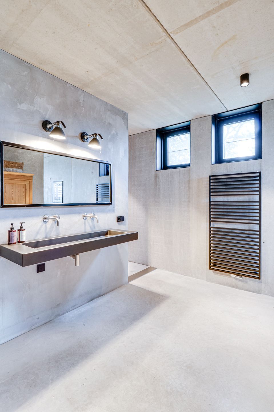 Minimalistische badkamer met wandgemonteerde wastafel, grote spiegel en twee kleine ramen boven een radiatorombouw.