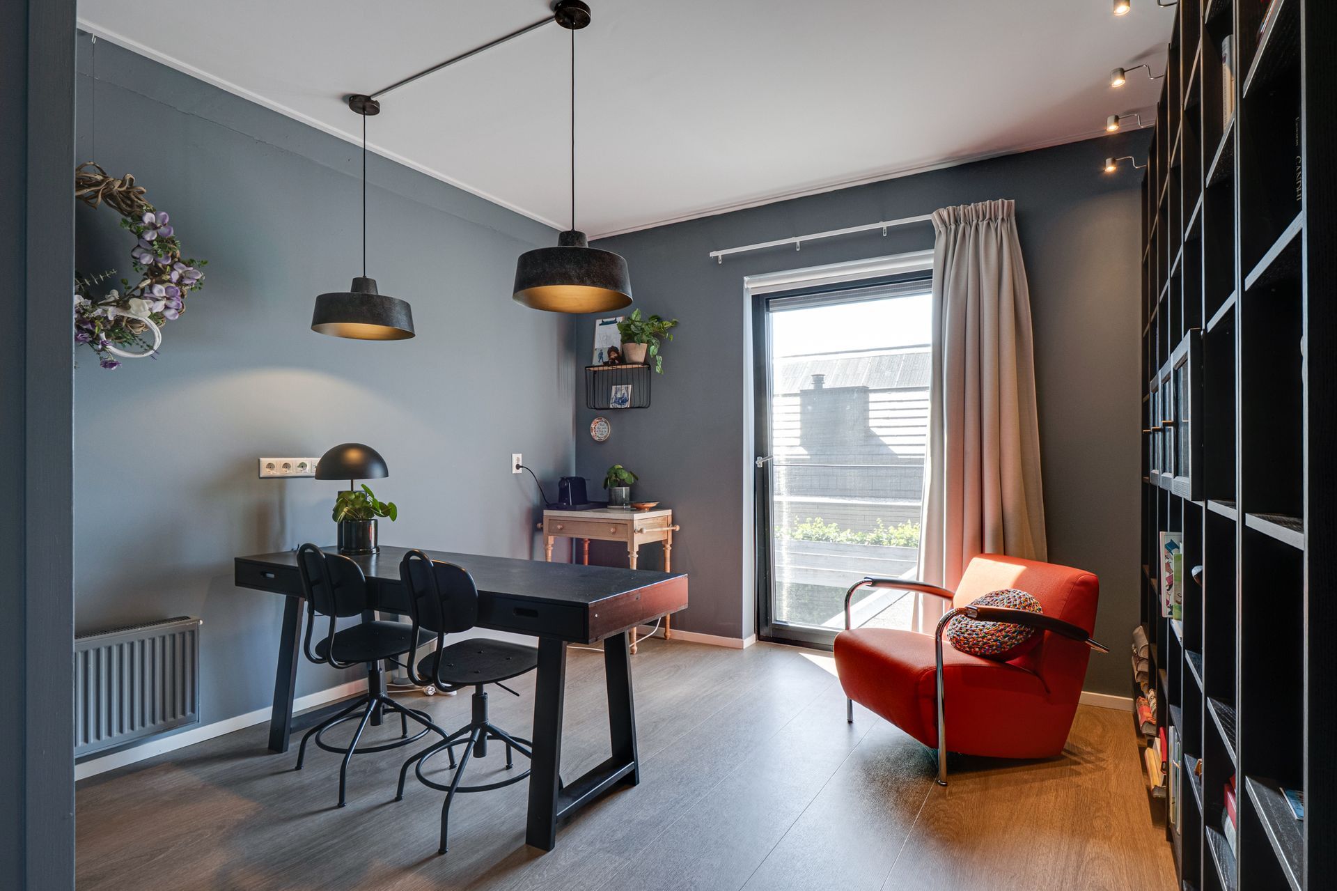Moderne thuiswerkplek met zwart bureau, hanglampen, rode stoel en groot raam.
