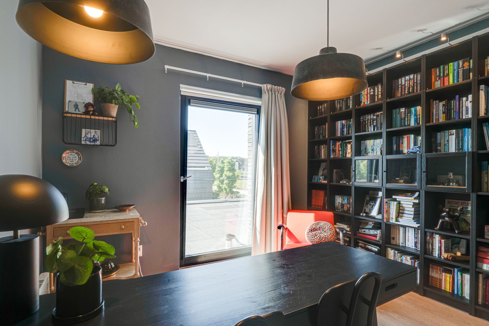 Moderne thuiswerkplek met zwart bureau, boekenkast aan de muur, hanglampen en een openslaande deur naar een licht balkon.