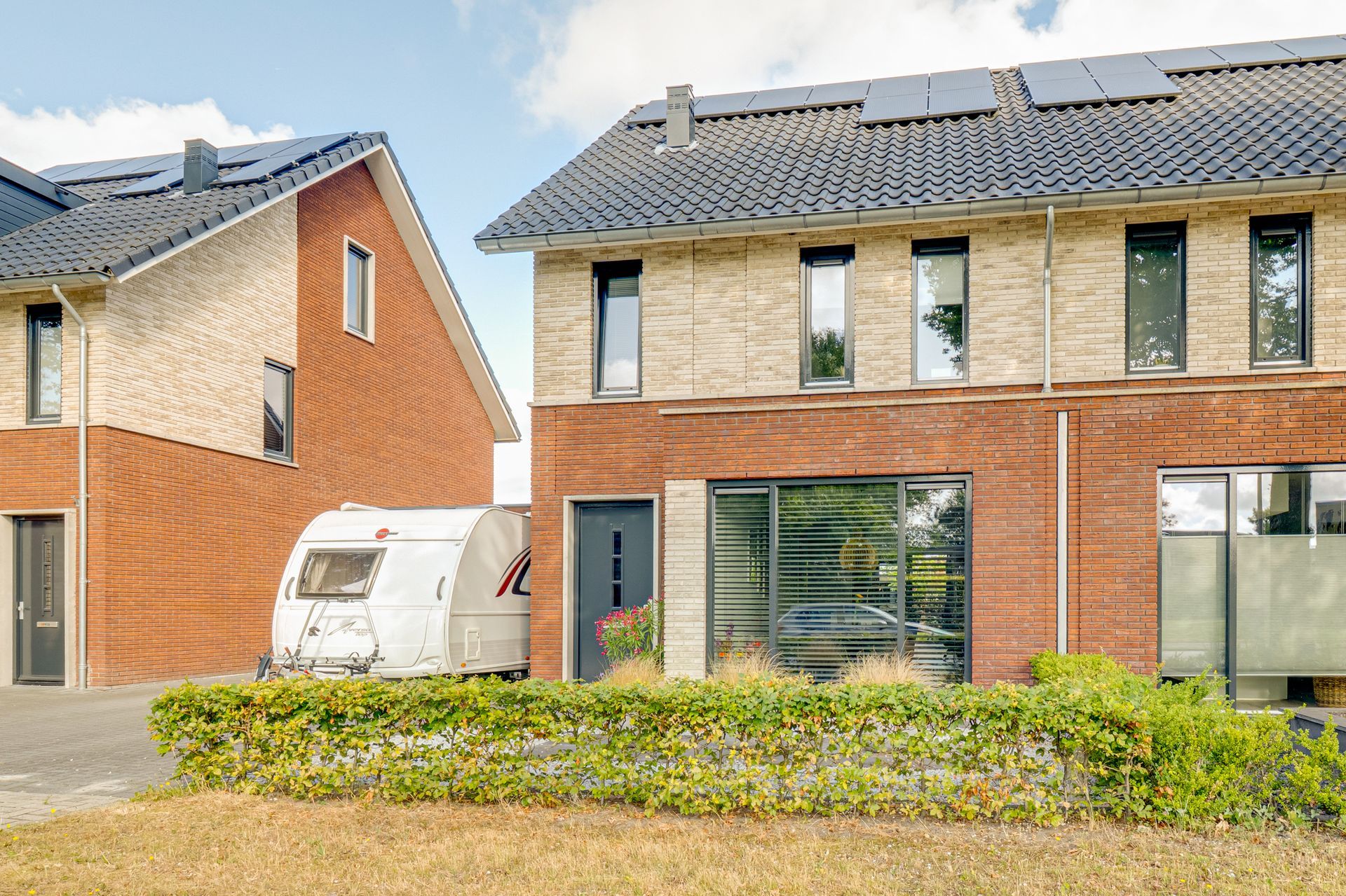 Moderne bakstenen rijtjeswoning met grote ramen, een heg en een geparkeerde witte caravan ervoor.