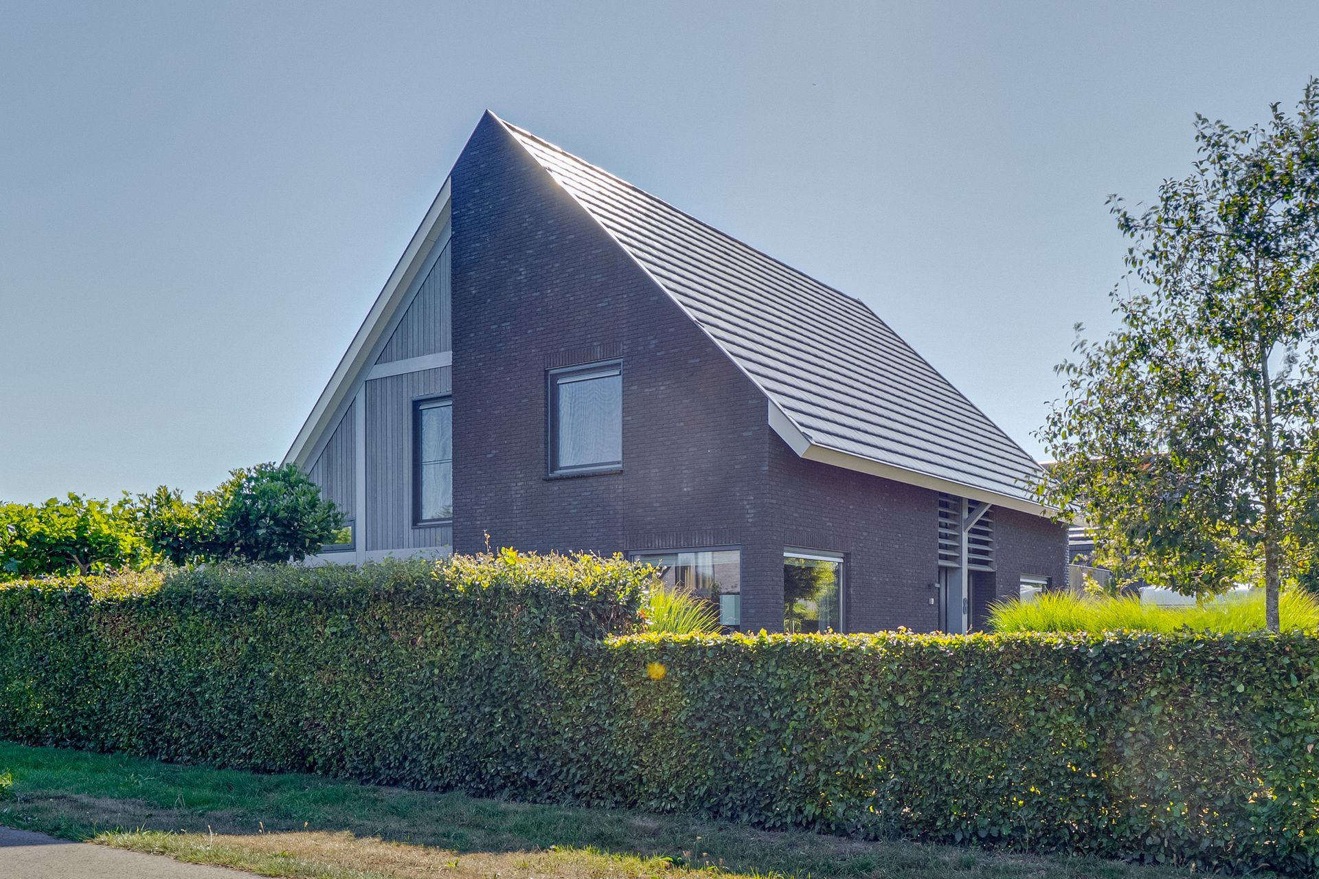 Moderne bakstenen woning met een steil dak, verscholen achter een strak gesnoeide heg, onder een heldere hemel.