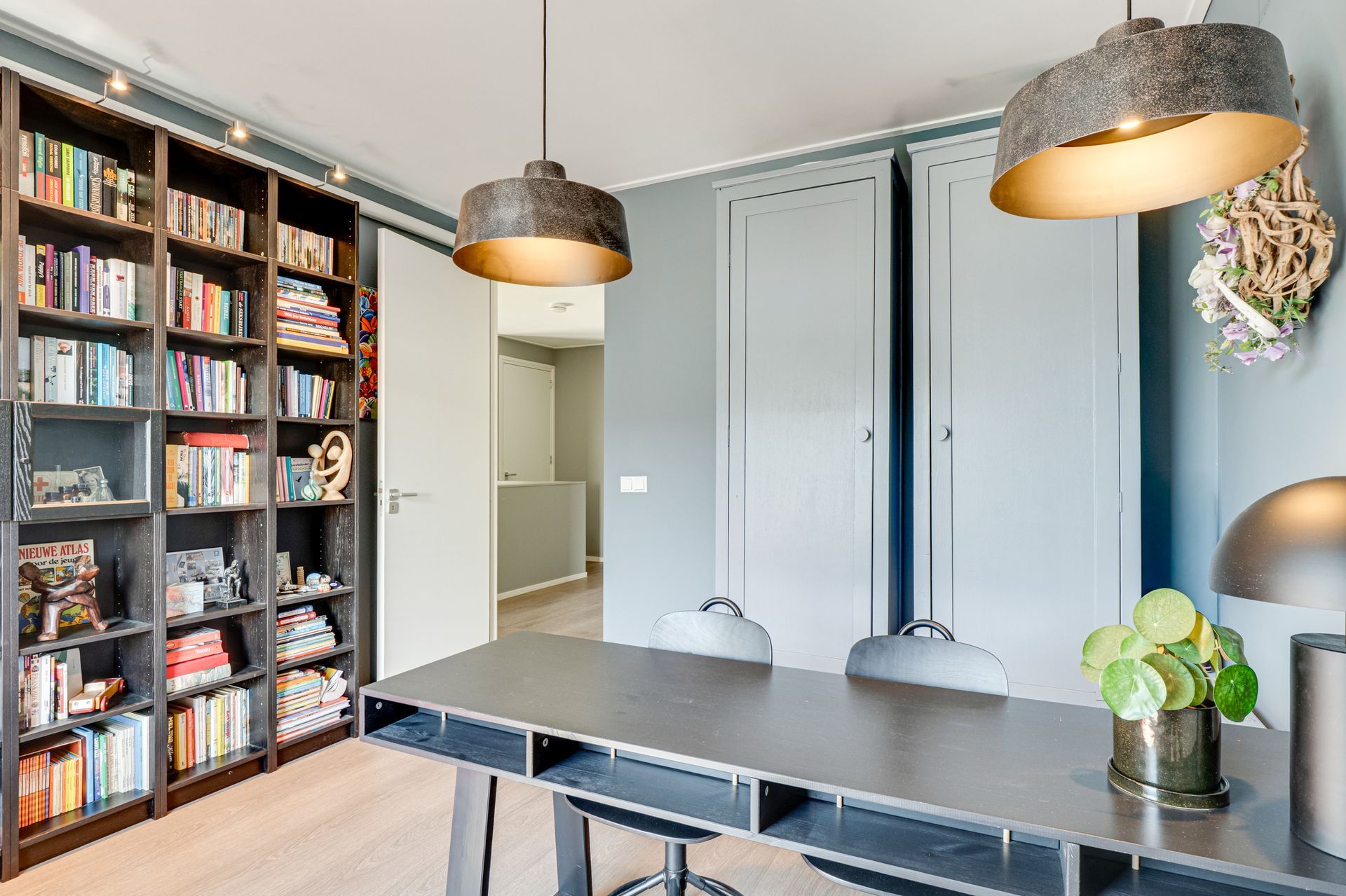 Moderne eetkamer met donkere tafel, hanglampen, boekenkast en grijze kastdeuren.