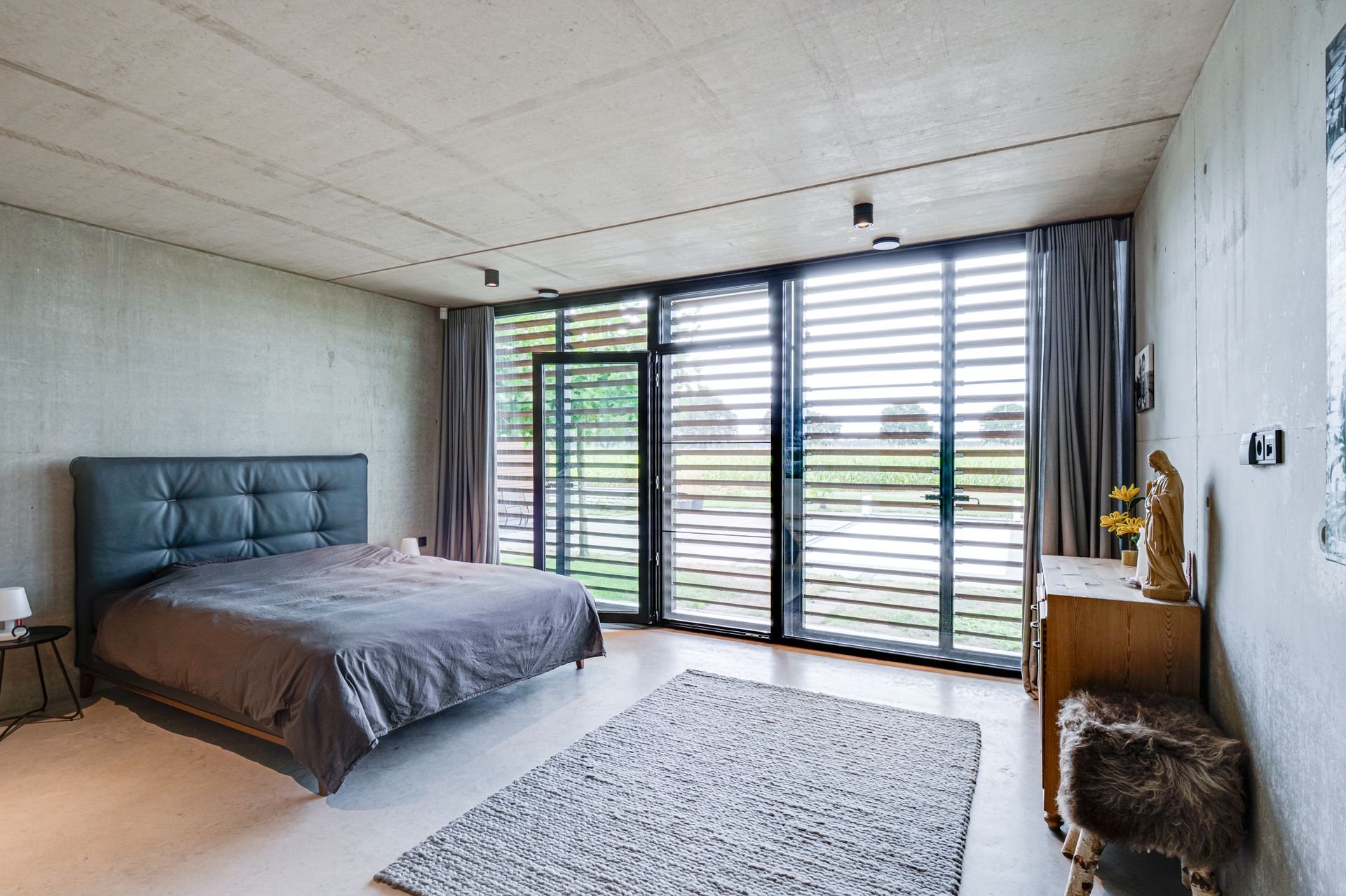 Moderne slaapkamer met een bed, vloerkleed en kamerhoge ramen met uitzicht op groen.