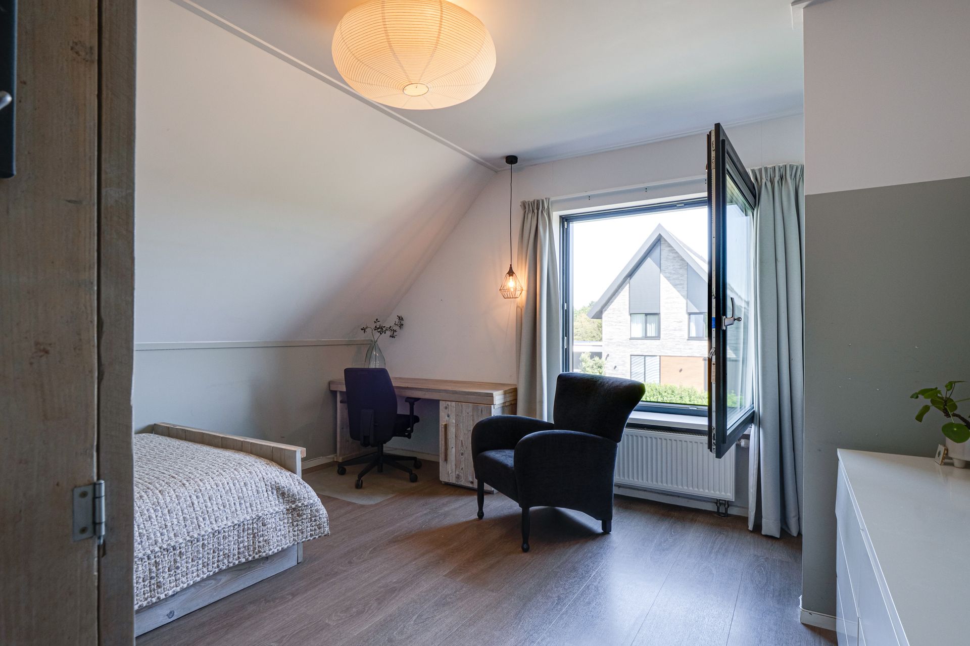Minimalistische slaapkamer met bed, bureau, stoel en een open raam in een lichte zolderkamer.