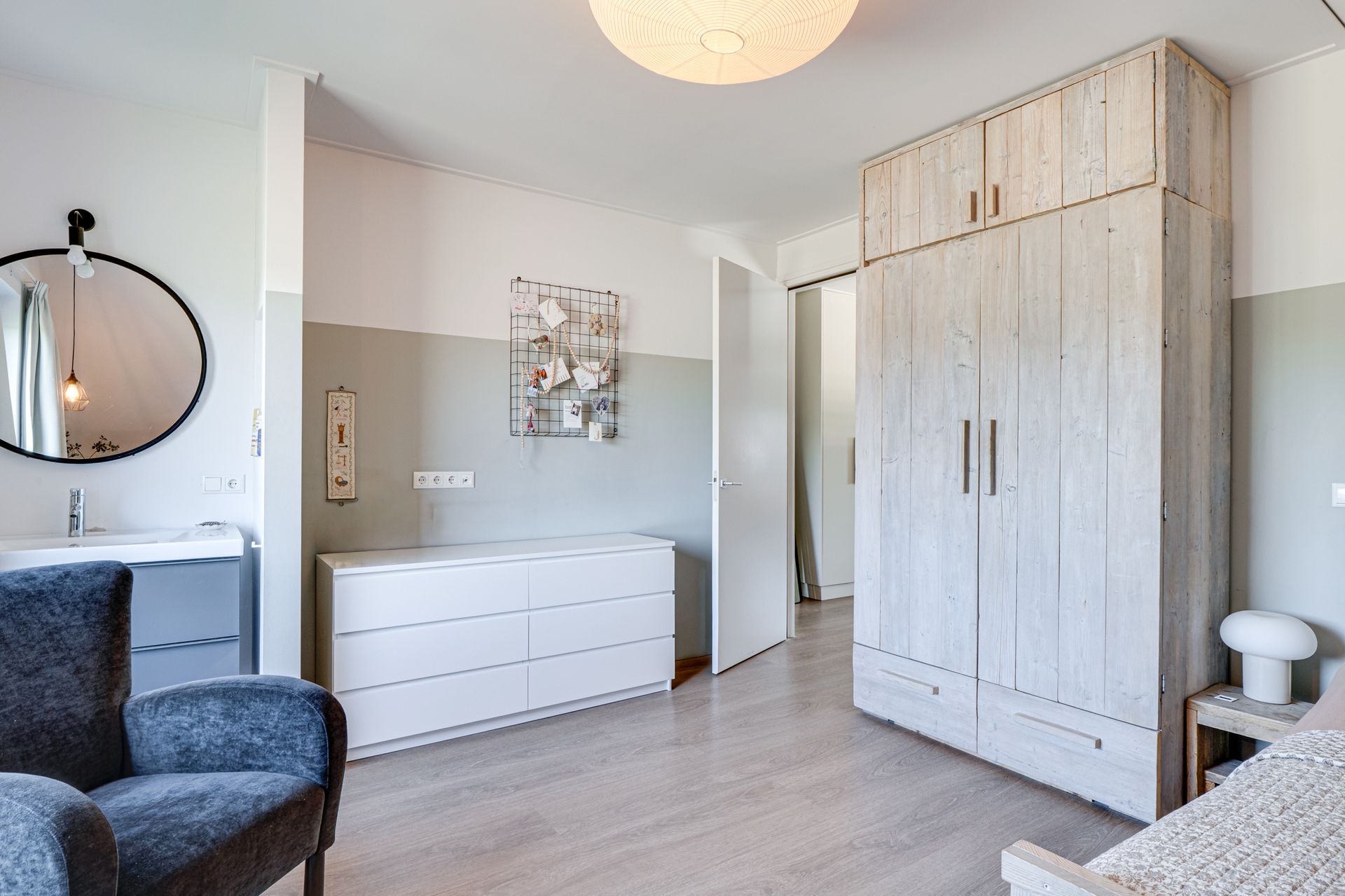 Lichte slaapkamer met witte commode, hoge kledingkast, kapspiegel en een blauwe stoel tegen lichtgrijze muren.