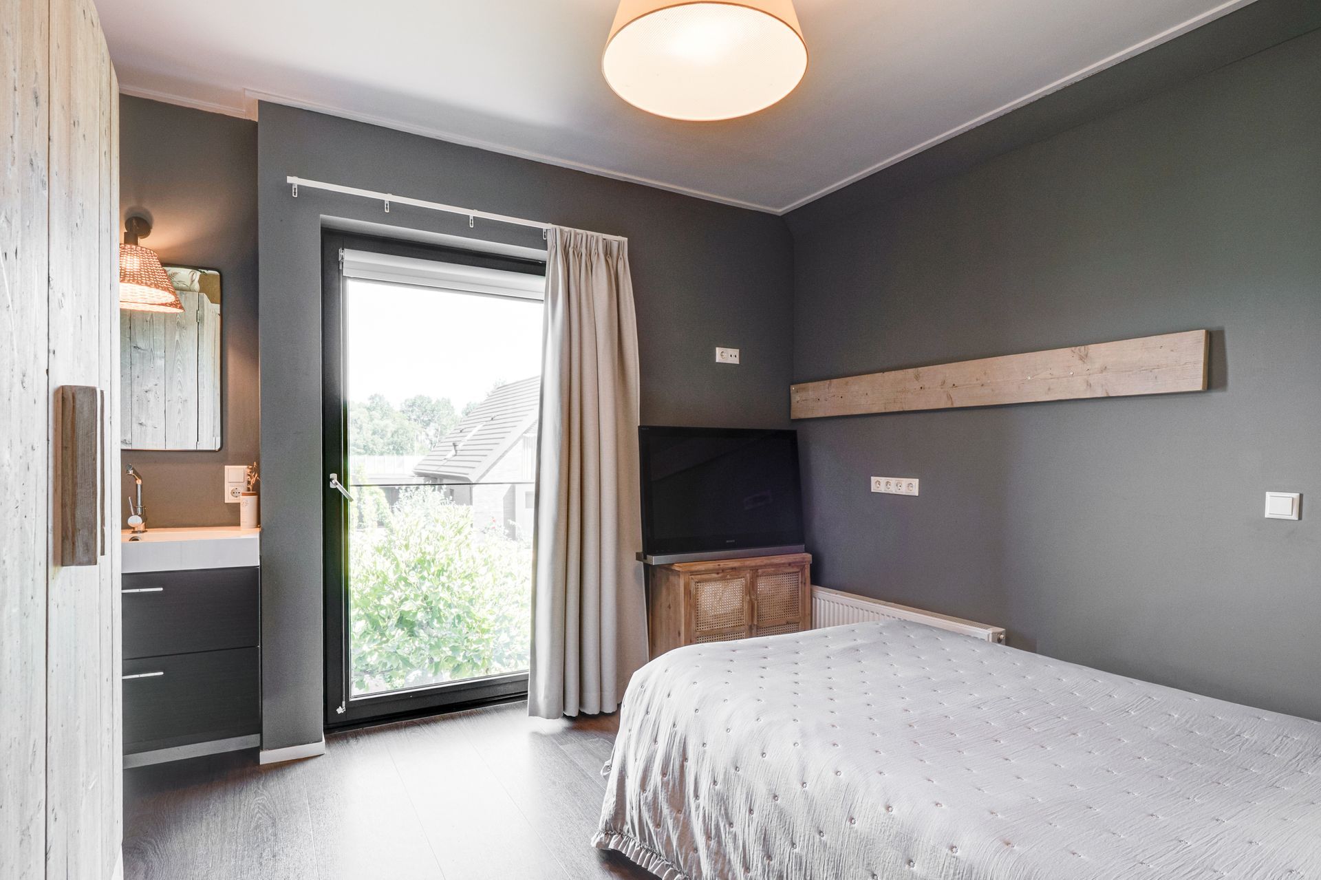 Moderne slaapkamer met grijze muren, bed, bureau en openslaande balkondeur met gordijnen.