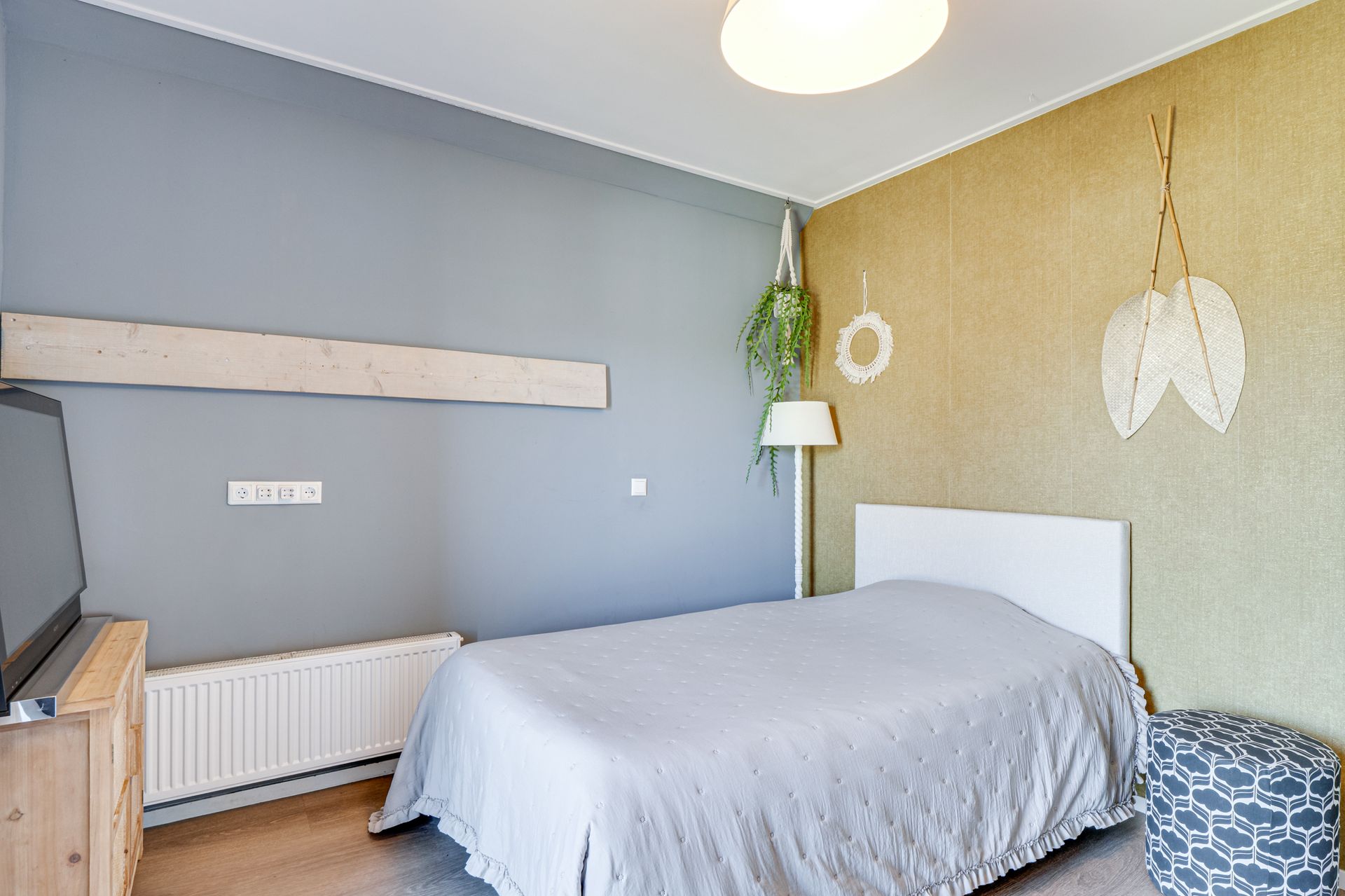 Kleine slaapkamer met grijze muren, een bed, radiator, tv en decoratieve hangplanten.