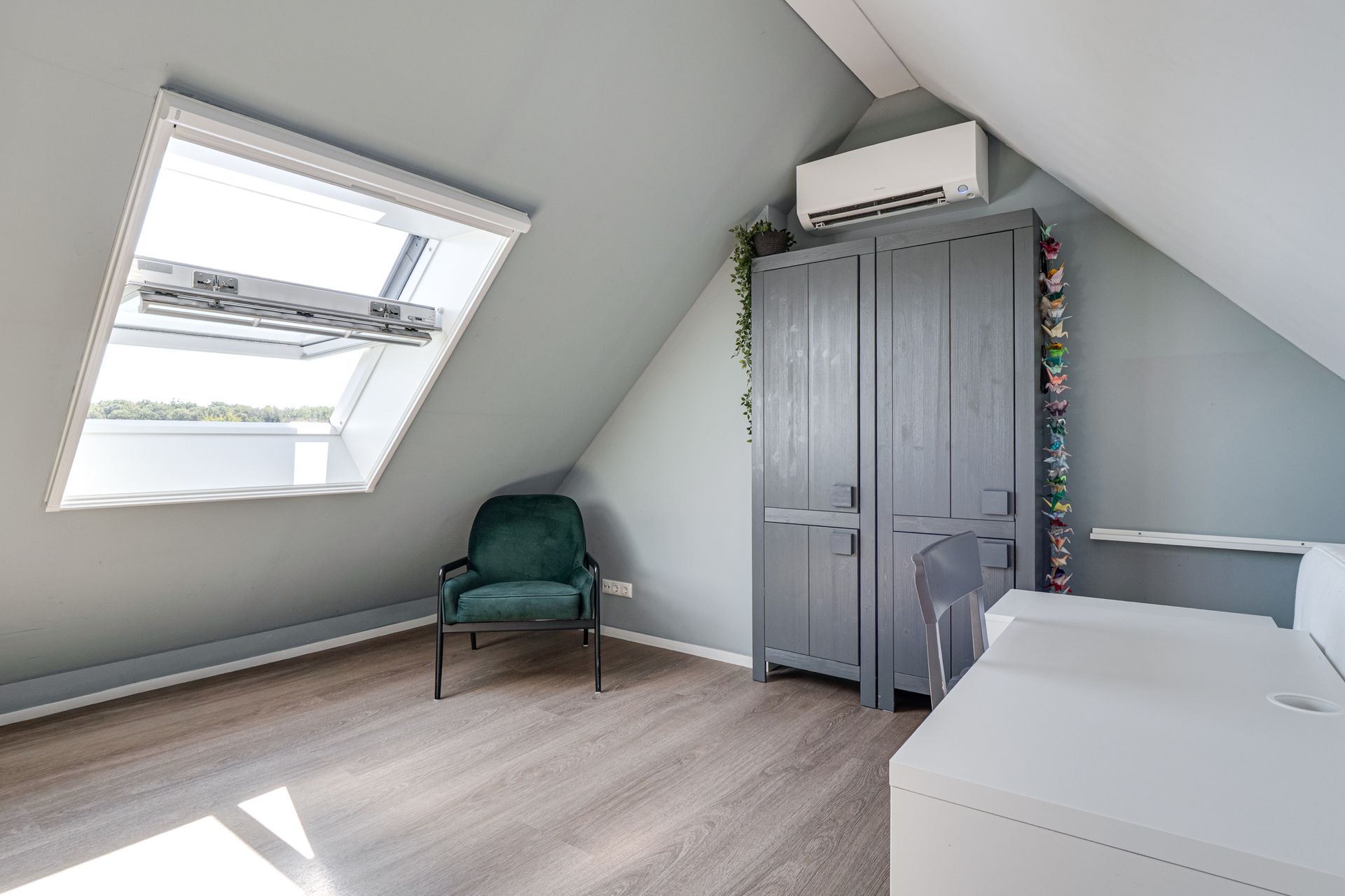 Zolderslaapkamer met dakraam, groene stoel, grijze kledingkast en een bed onder een schuin plafond.