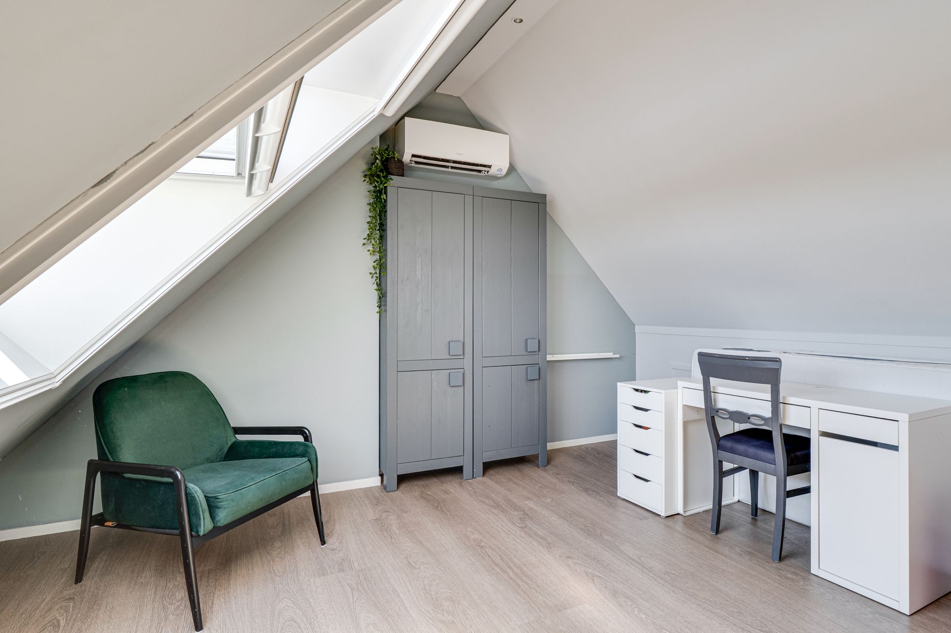 Zolderslaapkamer met groene stoel, grijze kledingkast en wit bureau onder een schuin plafond.