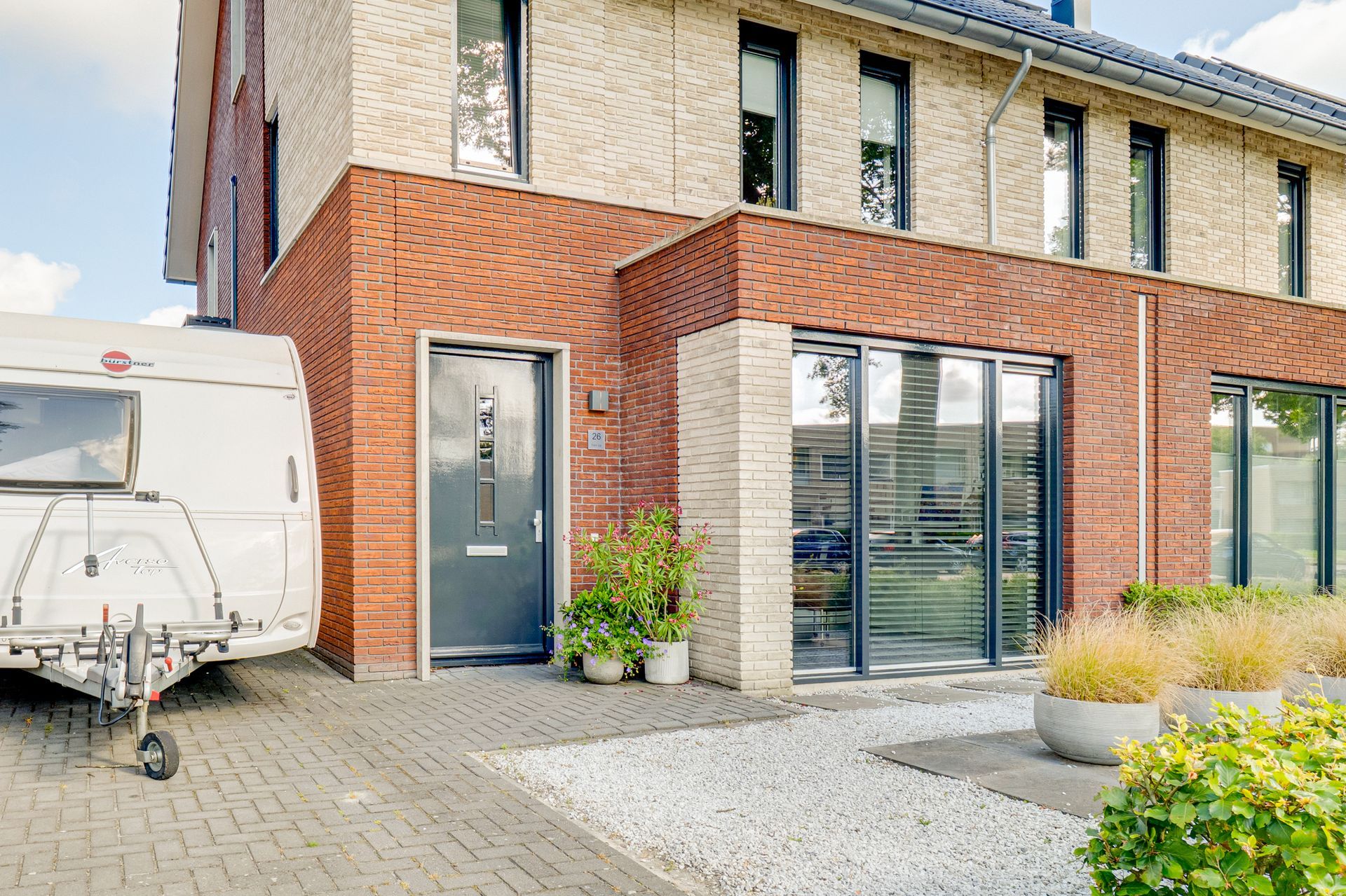 Moderne entree van een rijtjeshuis met grijze deur, grote ramen en een geparkeerde witte trailer naast een bakstenen gevel.
