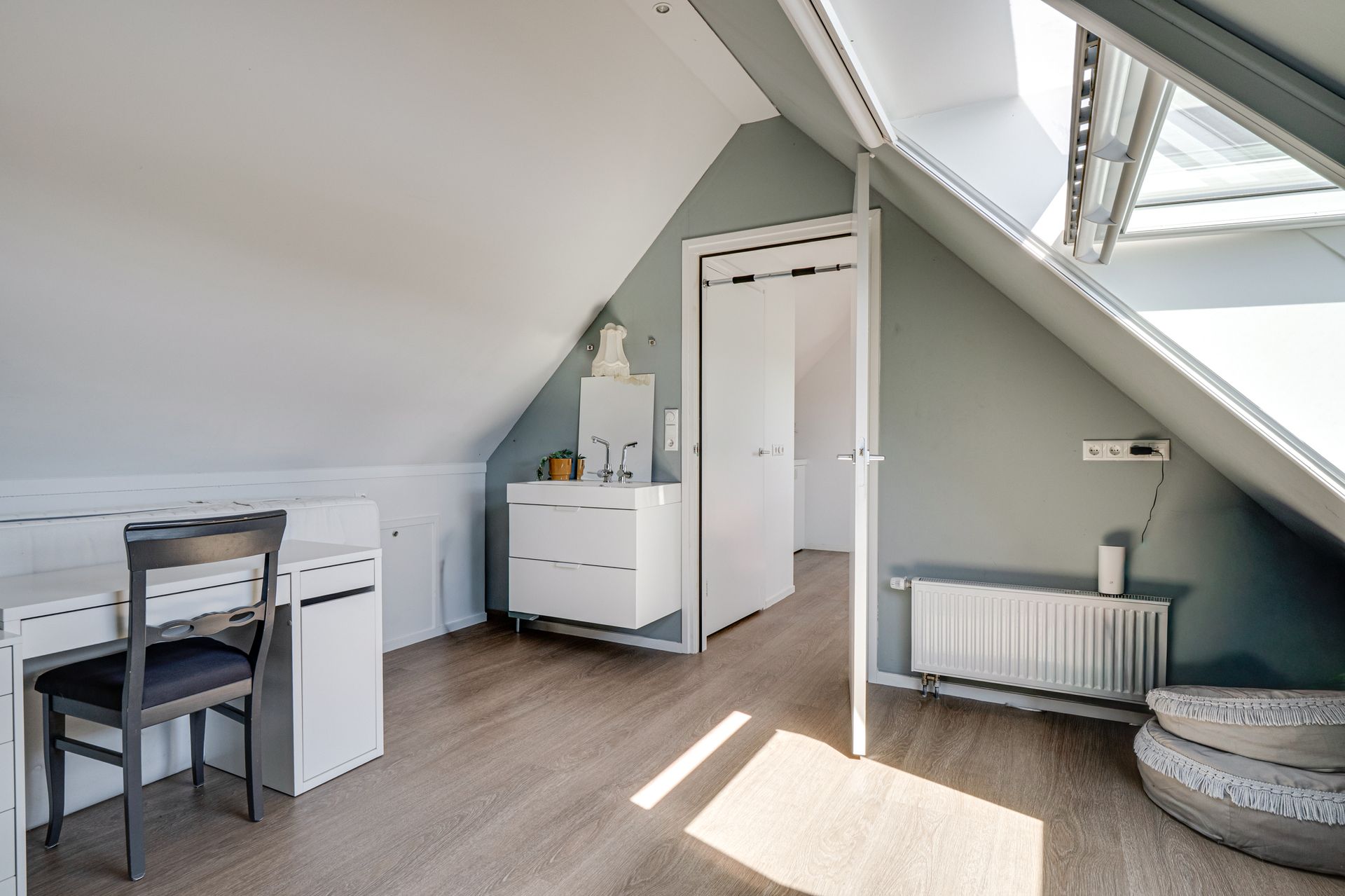 Kleine zolderkamer met bureau, witte commode, kledingkast, radiator en schuine dakramen.