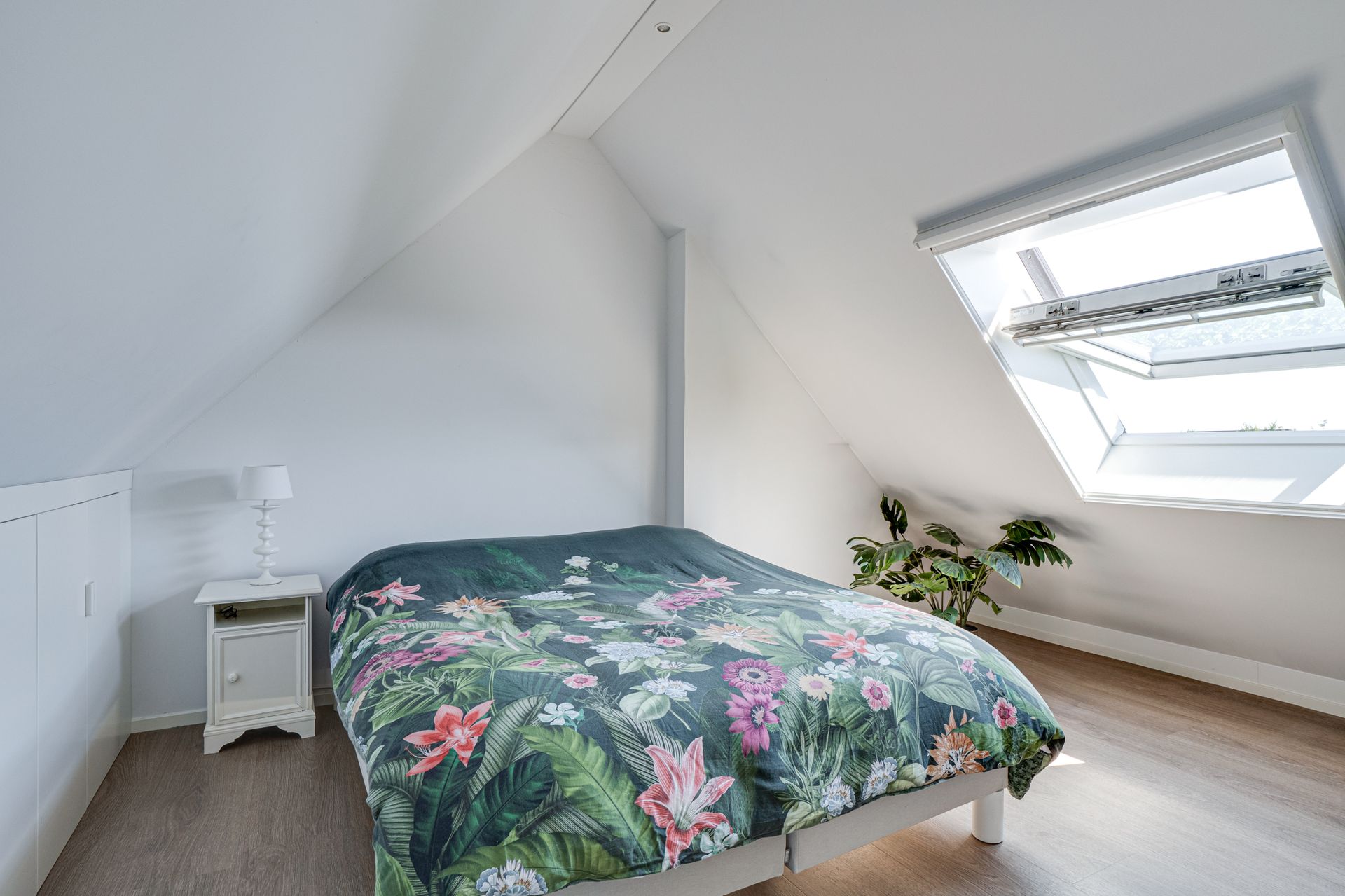 Lichte zolderkamer met een bloemensprei, witte muren, nachtkastje en dakraam.